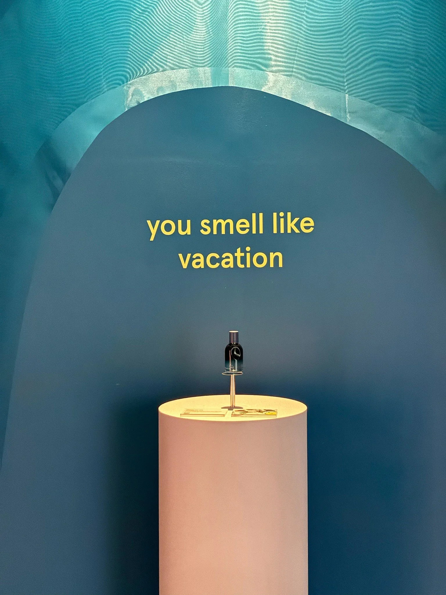 A bottle of perfume placed on a round pedestal with a blue background and the words 'you smell like vacation' written in yellow text.