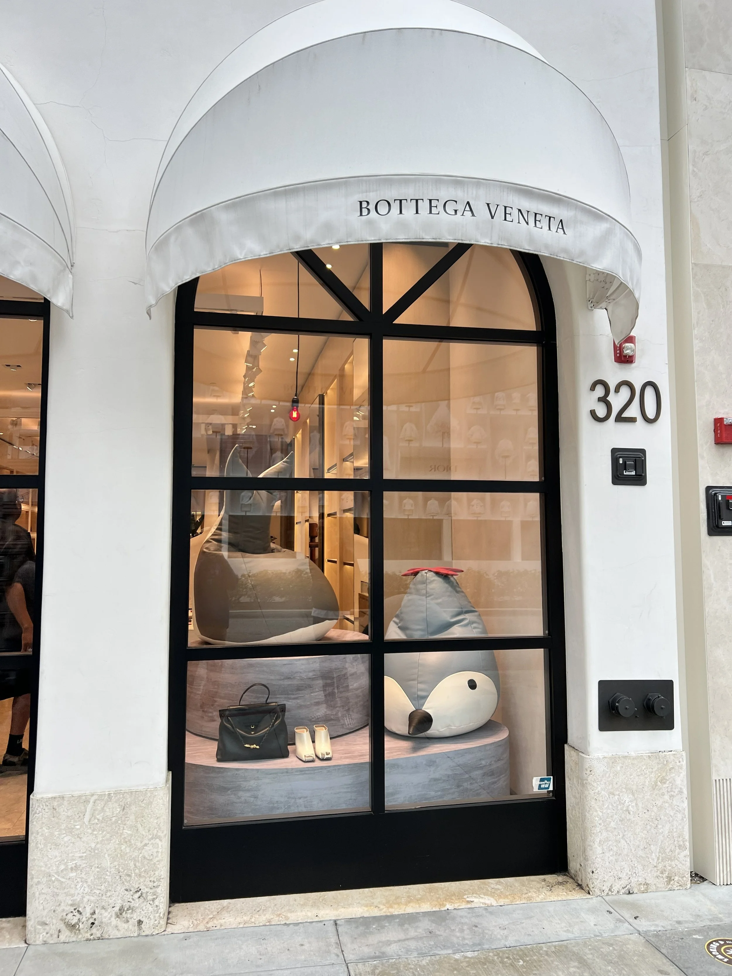 Storefront window display of Bottega Veneta with a large plush duck-shaped chair, a small handbag, two pairs of shoes, and other accessories inside.