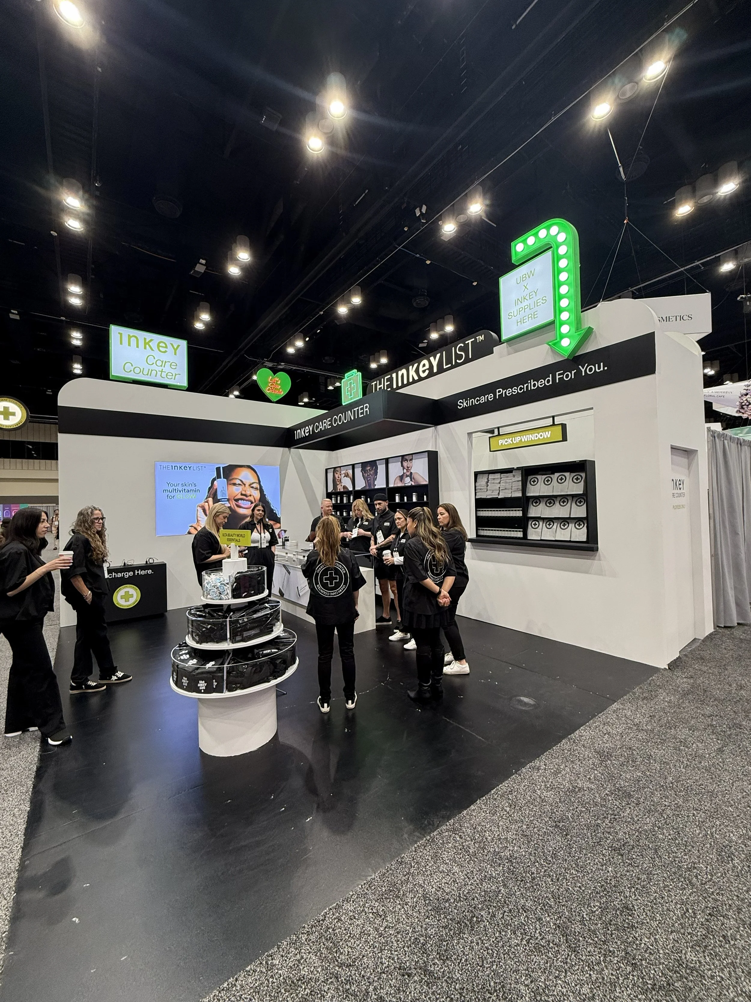 A trade show booth for Inkey Care Counter featuring staff members and product displays. The area has bright overhead lighting and signage, including a large digital screen displaying promotional content and a neon arrow sign pointing downward. Severa