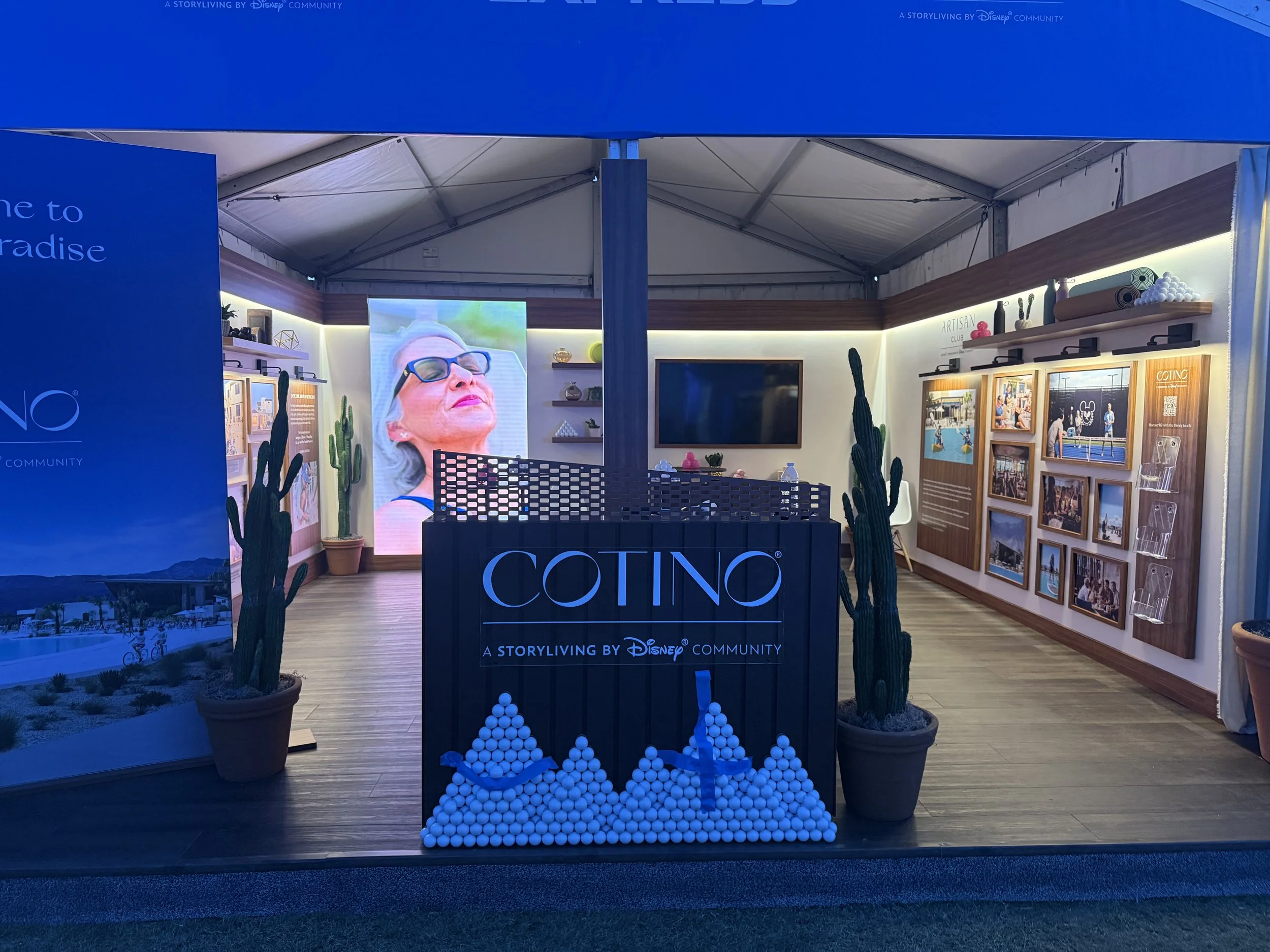 An indoor exhibition booth with blue walls and a large screen displaying a woman's face wearing sunglasses, surrounded by potted cacti and shelves with decorative items, featuring the branding 'COTINO' and the phrase 'A STORYLIVING BY Disney COMMUNIT