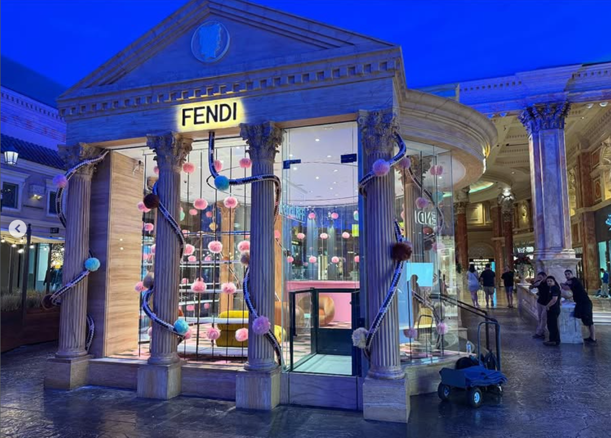 Decorative store facade designed to look like a classical building with columns and a pediment, labeled 'FENDI'. It features colorful pom-pom decorations and a climbing structure inside, inside a shopping mall.
