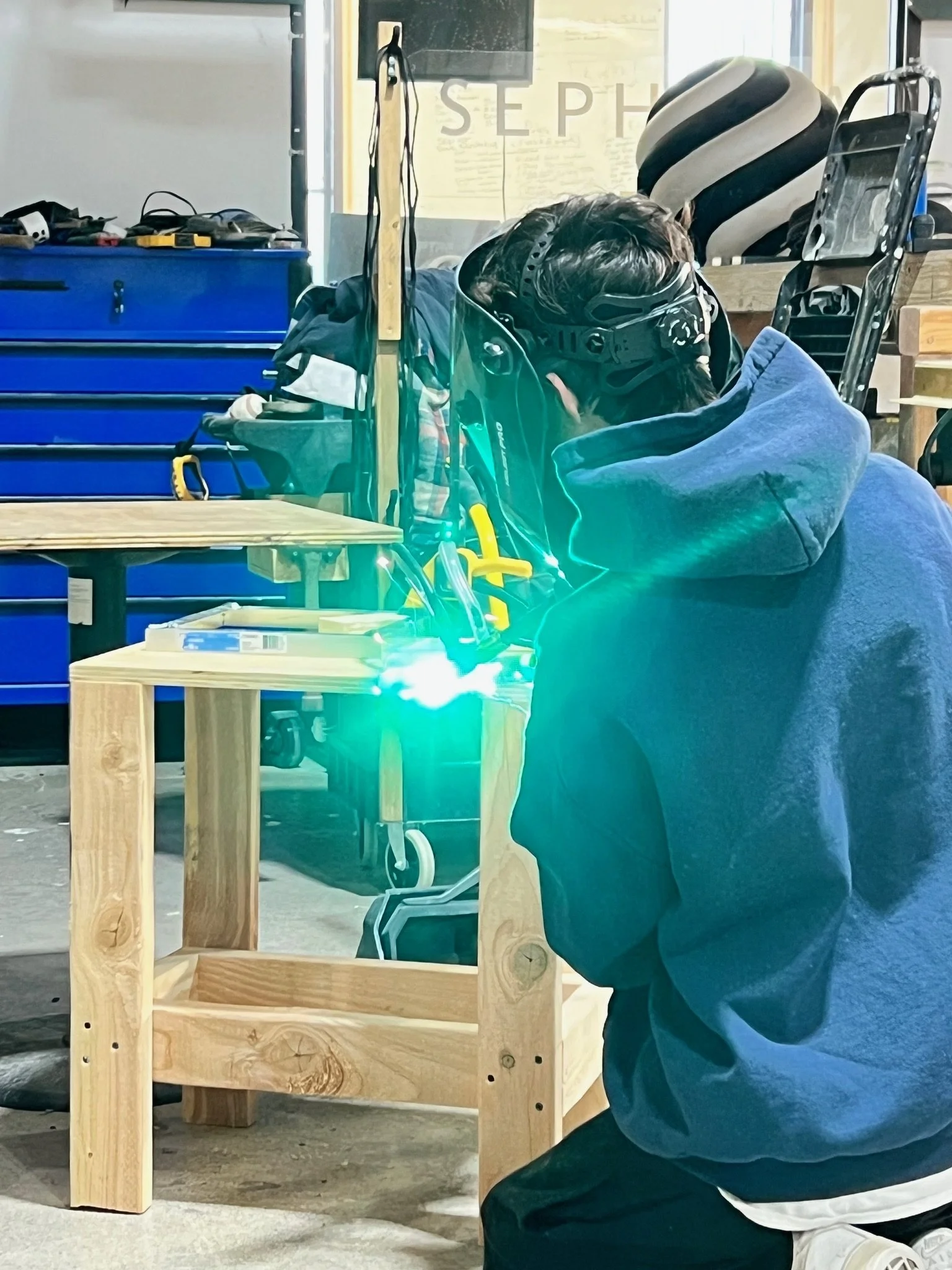 Person welding a wooden table in a workshop, wearing a welding helmet and protective gear.