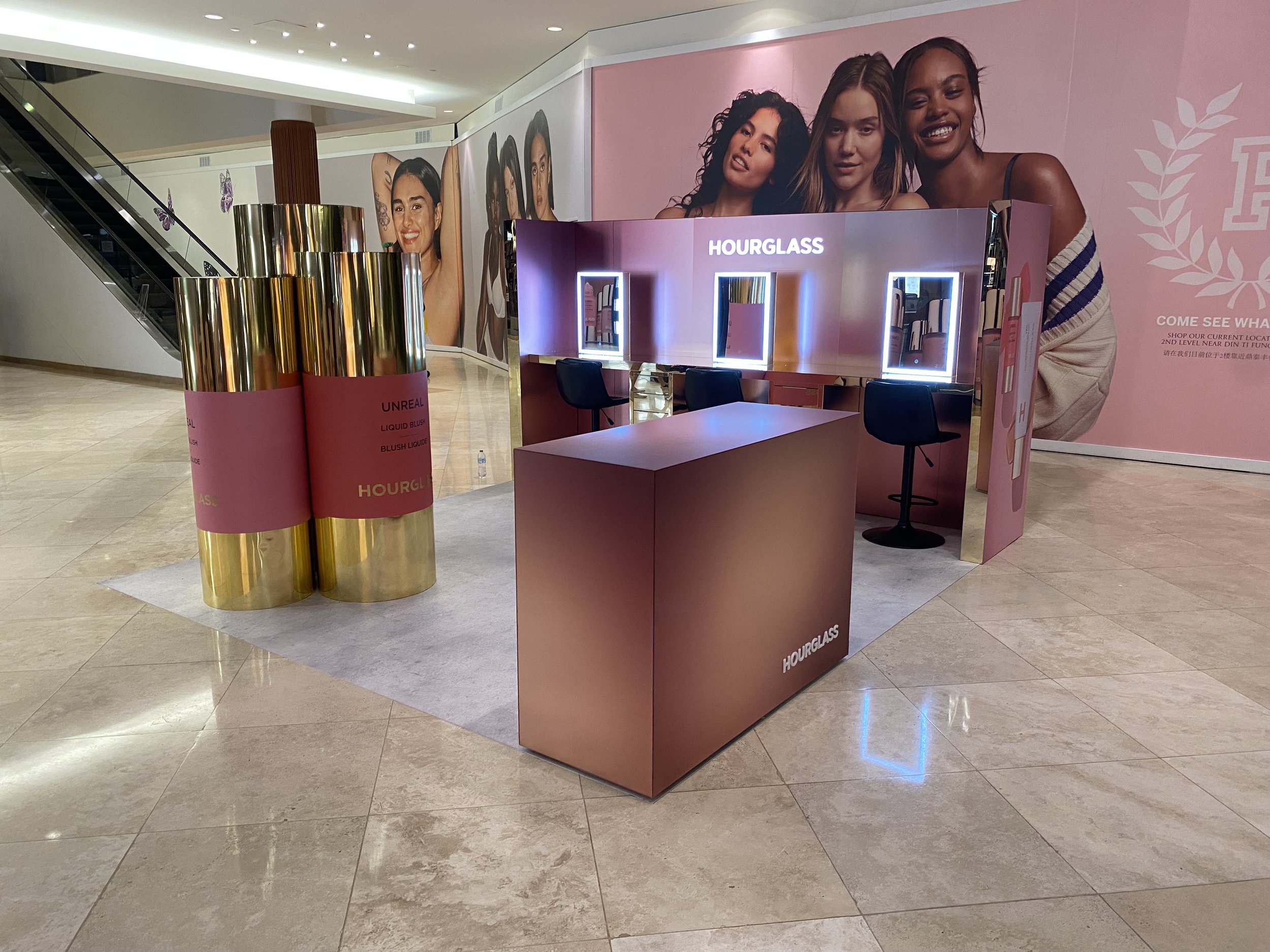 An indoor promotional display for Hourglass cosmetics, featuring large posters of women with diverse skin tones and makeup looks. There are illuminated mirrors and a table with three black chairs, set against a pink background with brand branding.