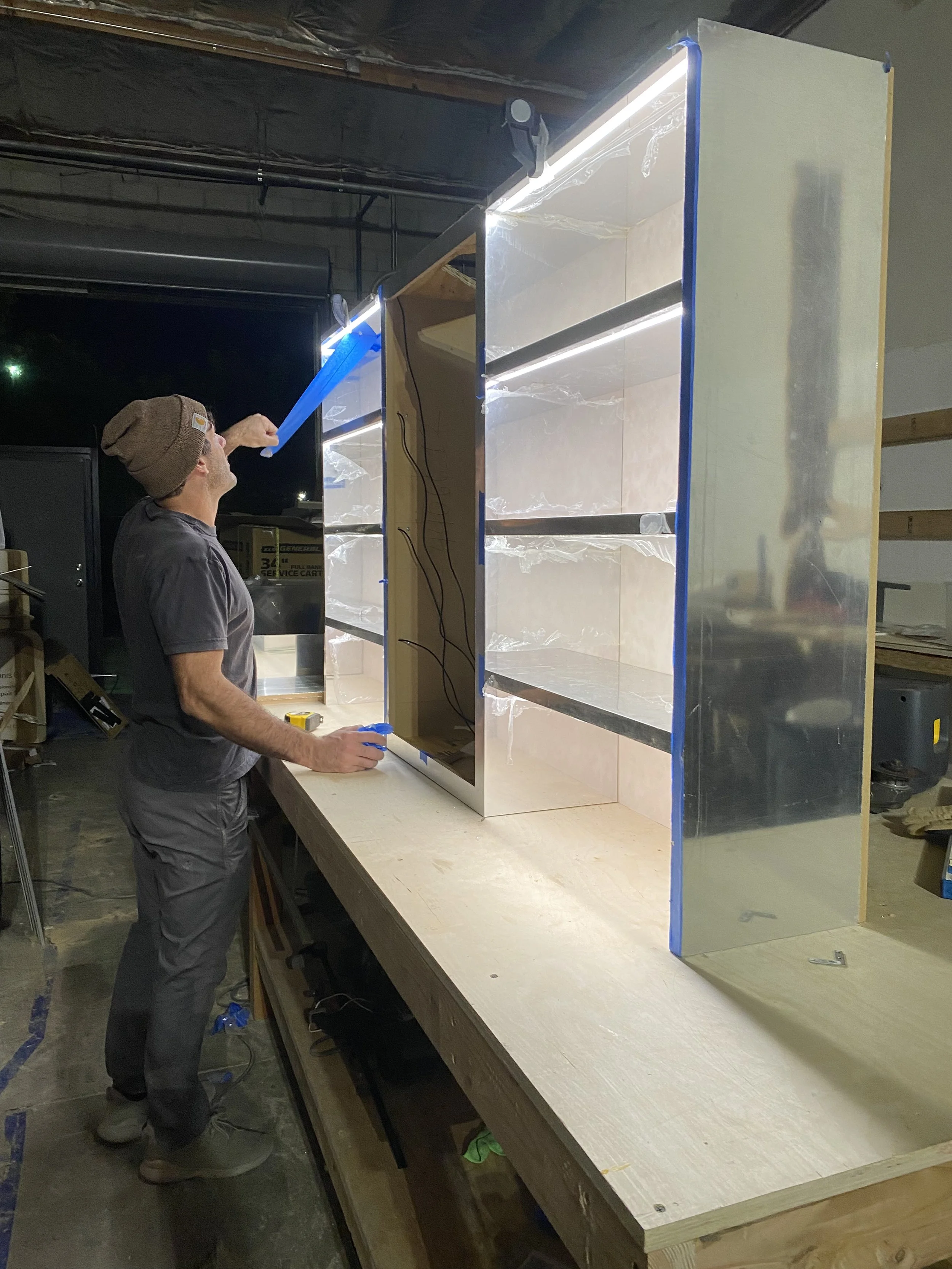 A man wearing a brown beanie and gray t-shirt is working on a large, partially assembled display or cabinet with glass and metal shelving in a workshop at night.
