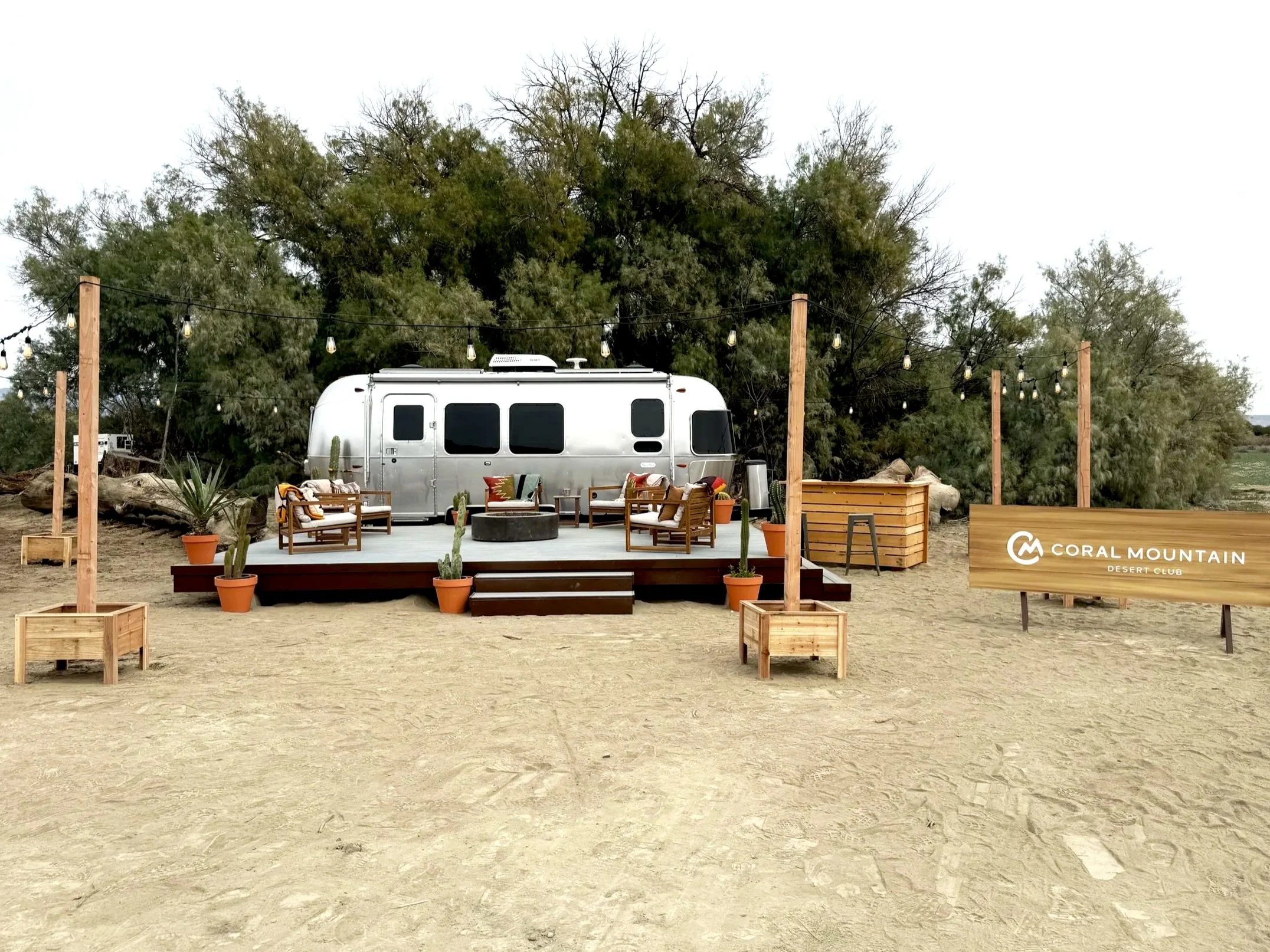 A venue with a wooden stage, outdoor seating, string lights, potted plants, and a sign that reads 'Coral Mountain Desert Club' with a trailer in the background and trees behind.