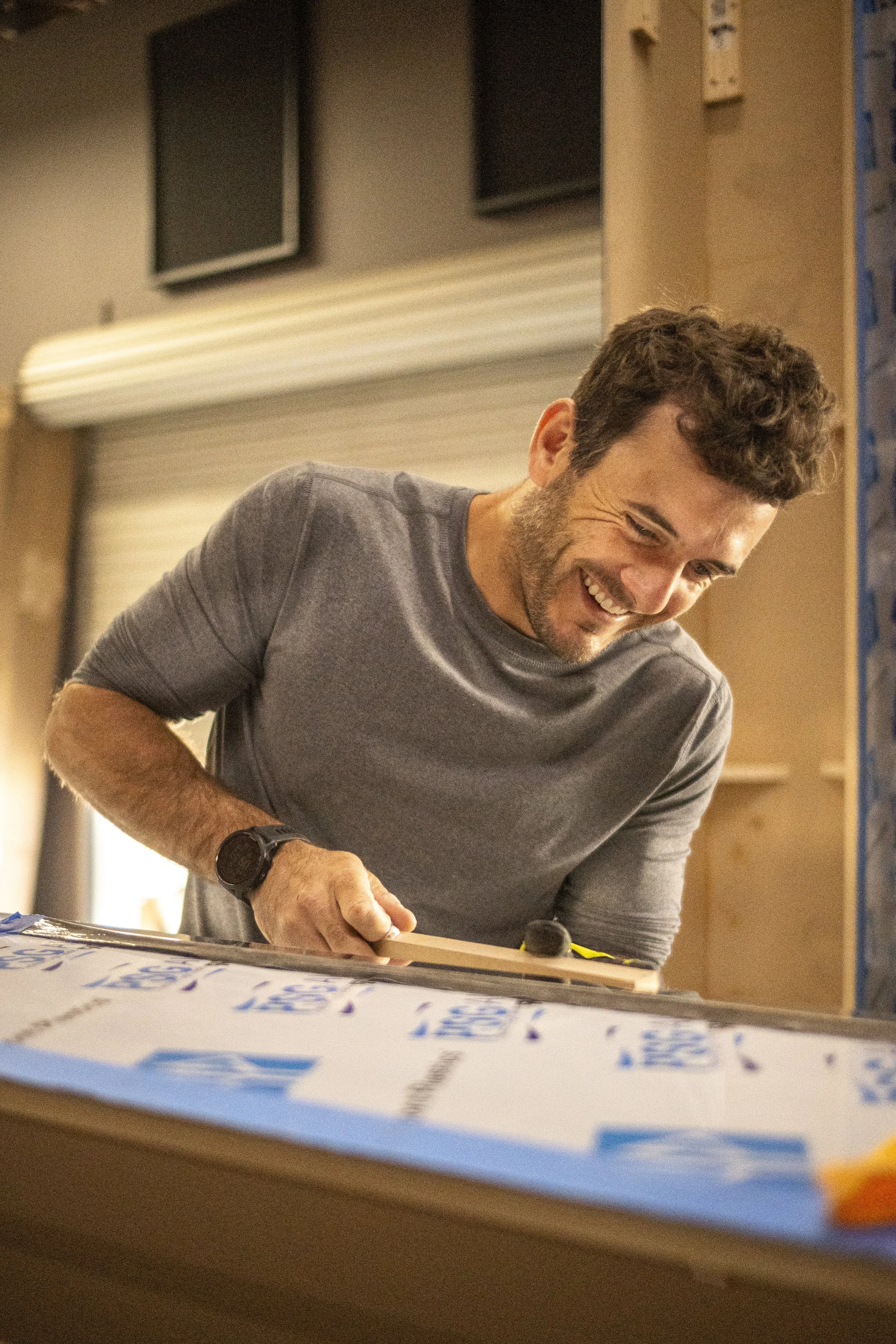 A man with curly hair and a beard, wearing a gray t-shirt and a black watch, smiling while writing on a large piece of paper on a table in a room with beige walls and black wall-mounted monitors.