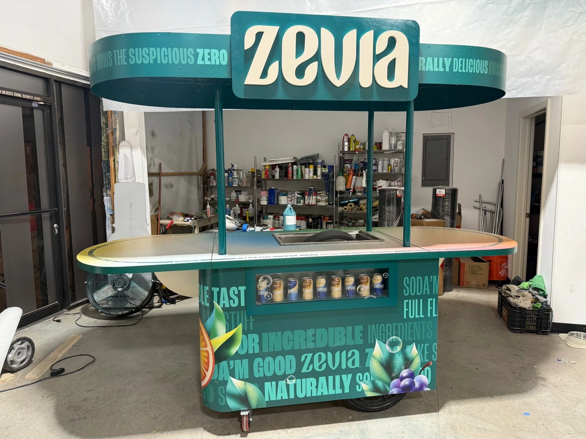 A beverage kiosk with a teal rounded top sign and a front panel displaying cans of Zevia soda in a vending area. The sign and panel highlight Zevia's natural ingredients and zero sugar content. The background shows shelves with supplies and some misc