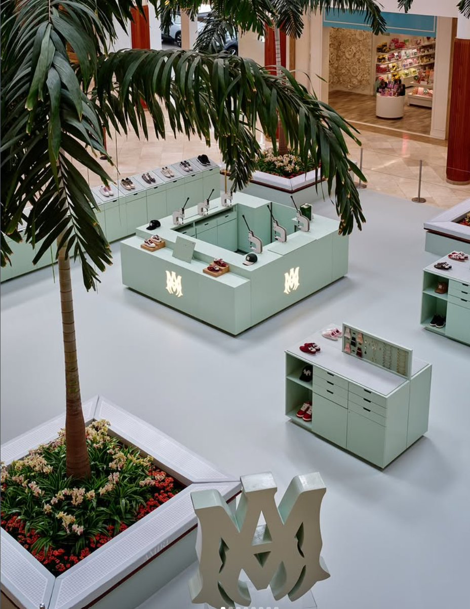 Empty retail kiosk with shoes on display in a mall, surrounded by plants and flower bed, with store entrance in background.