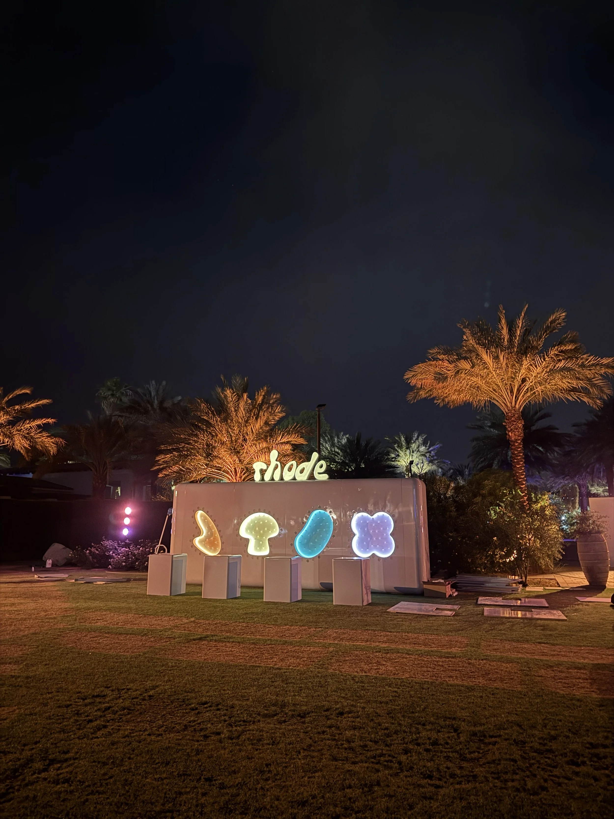 Night scene with palm trees illuminated by lights, a sign that says 'shade', and colorful illuminated shapes on display on a white panel, set in a landscaped area.
