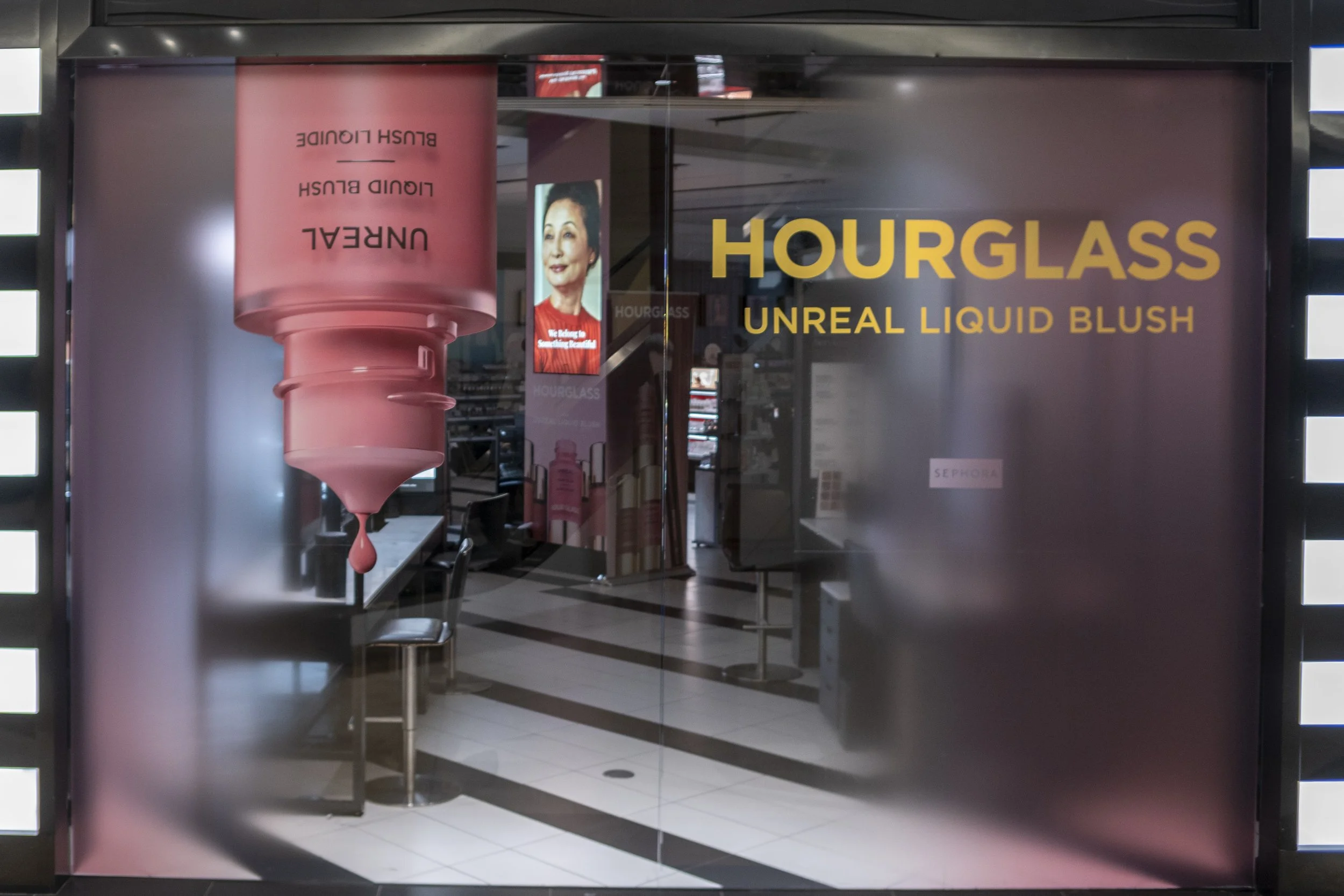 Store window display with large pink container of blush and text promoting Hourglass Unreal Liquid Blush, with interior view of mall or shopping center behind.