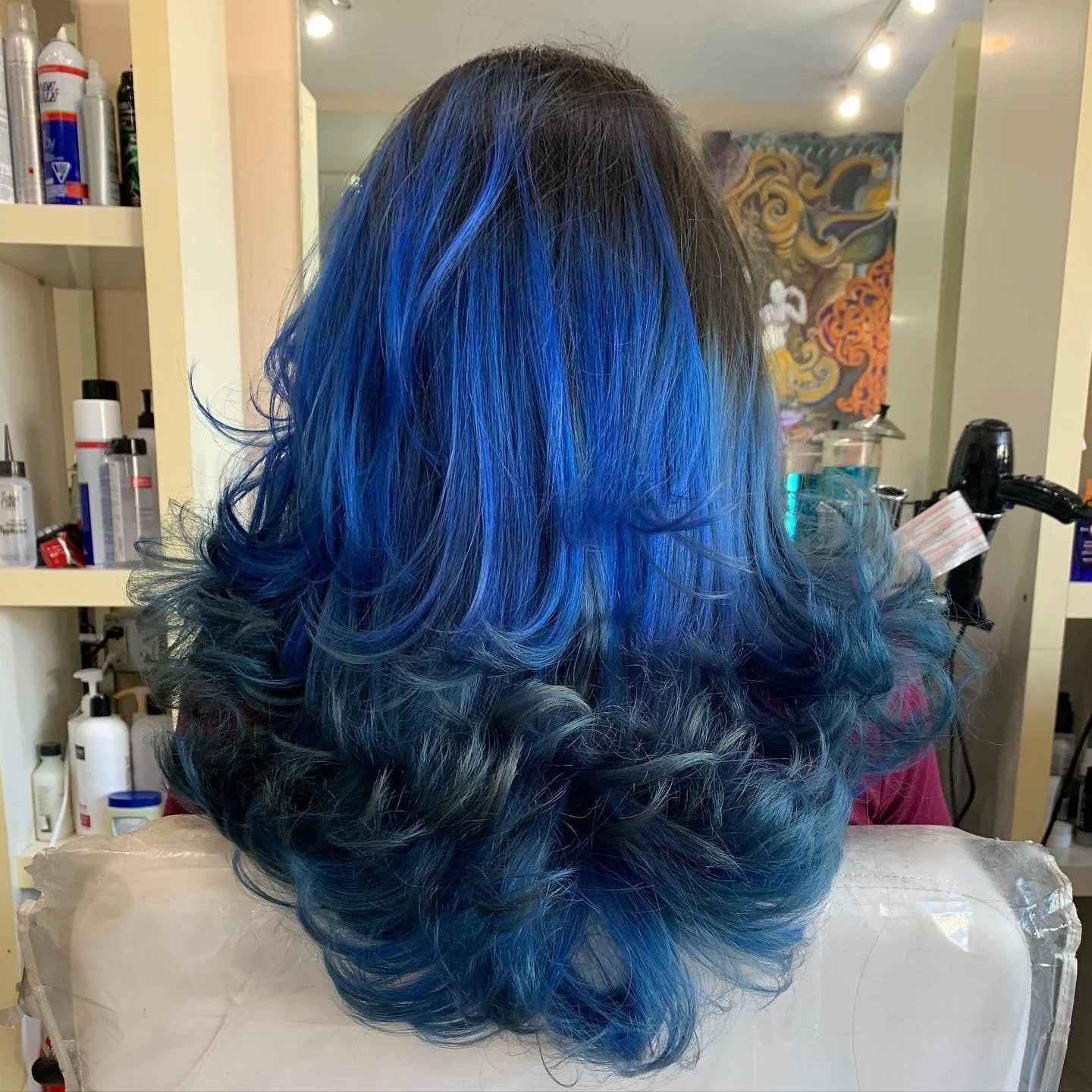 A person's hair dyed with shades of blue, teal, and black styled in loose curls, seen from the back in a salon setting.