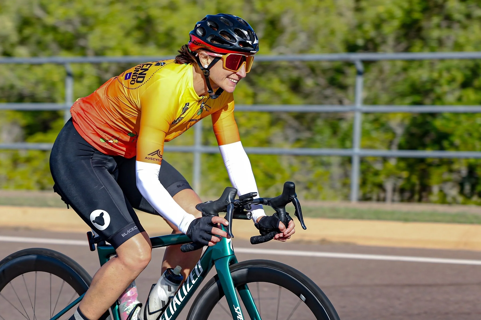 A female cyclist wearing a yellow and orange cycling jersey, black shorts, a black helmet, and sunglasses riding a teal Specialized road bike on a paved path with greenery in the background.