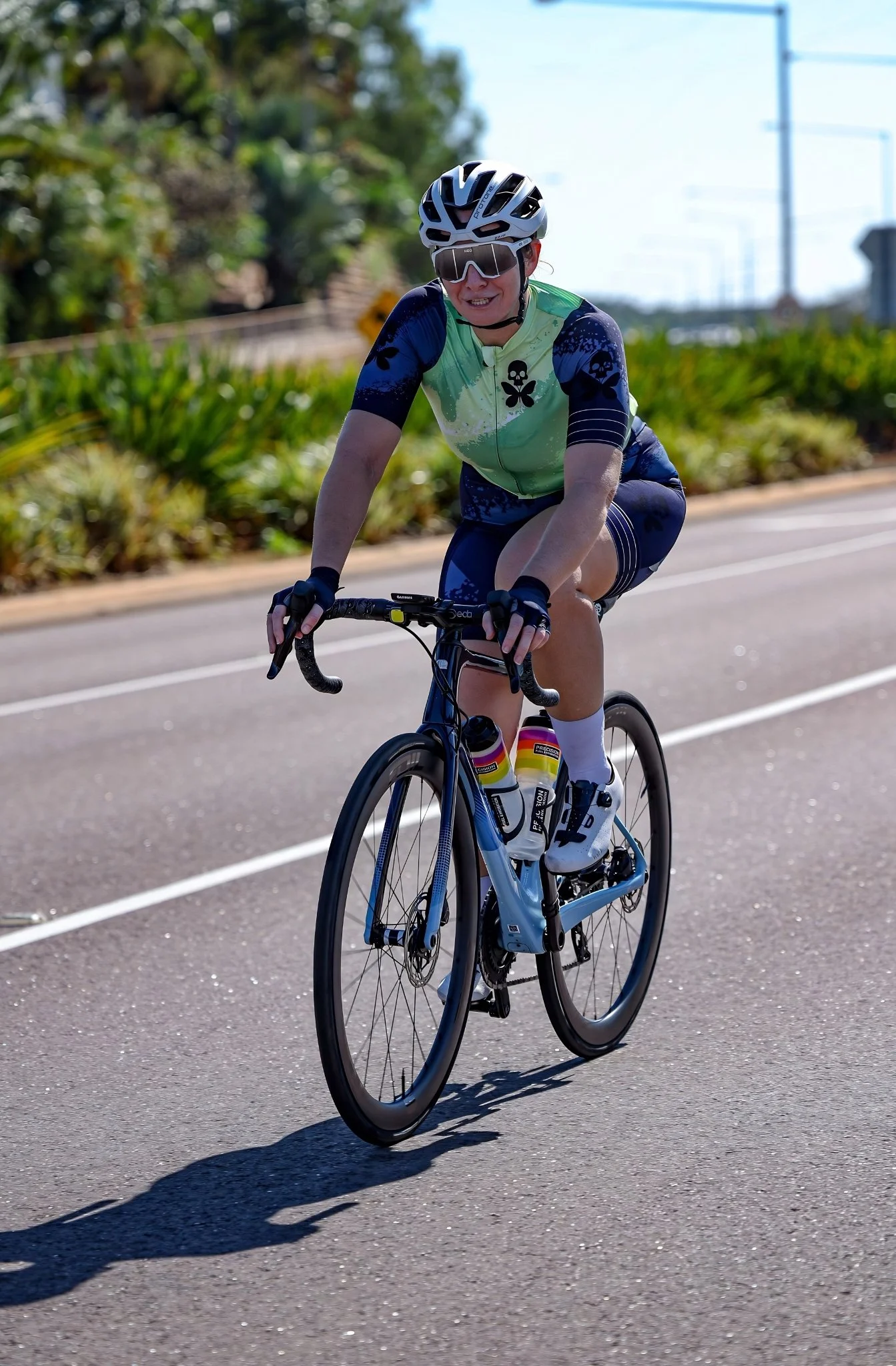 A woman riding a road bike on a paved path, wearing a white helmet, sunglasses, and a colorful cycling jersey with matching shorts.