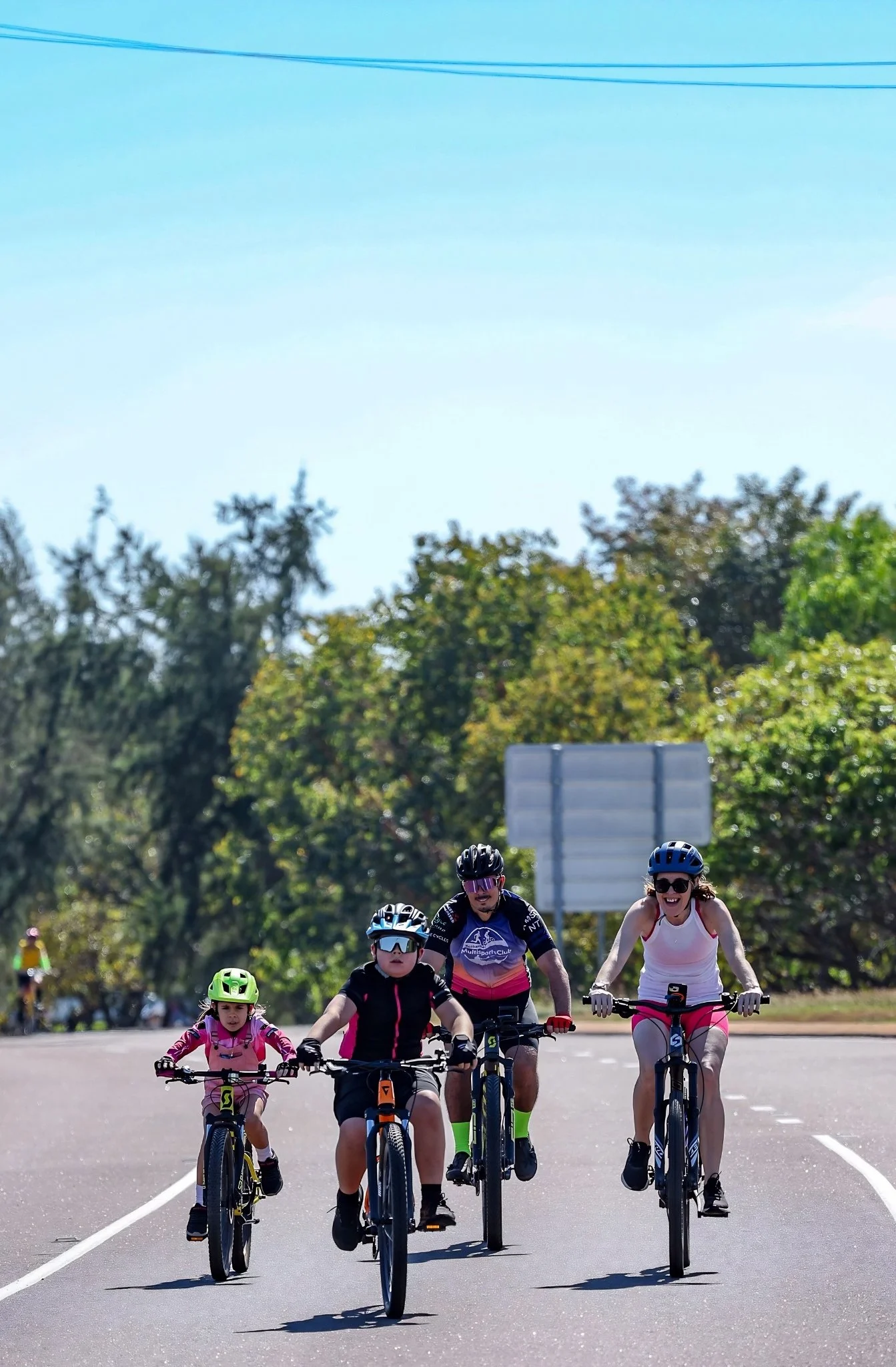 A group of four people riding bikes together on a sunny day, with greenery and trees in the background.