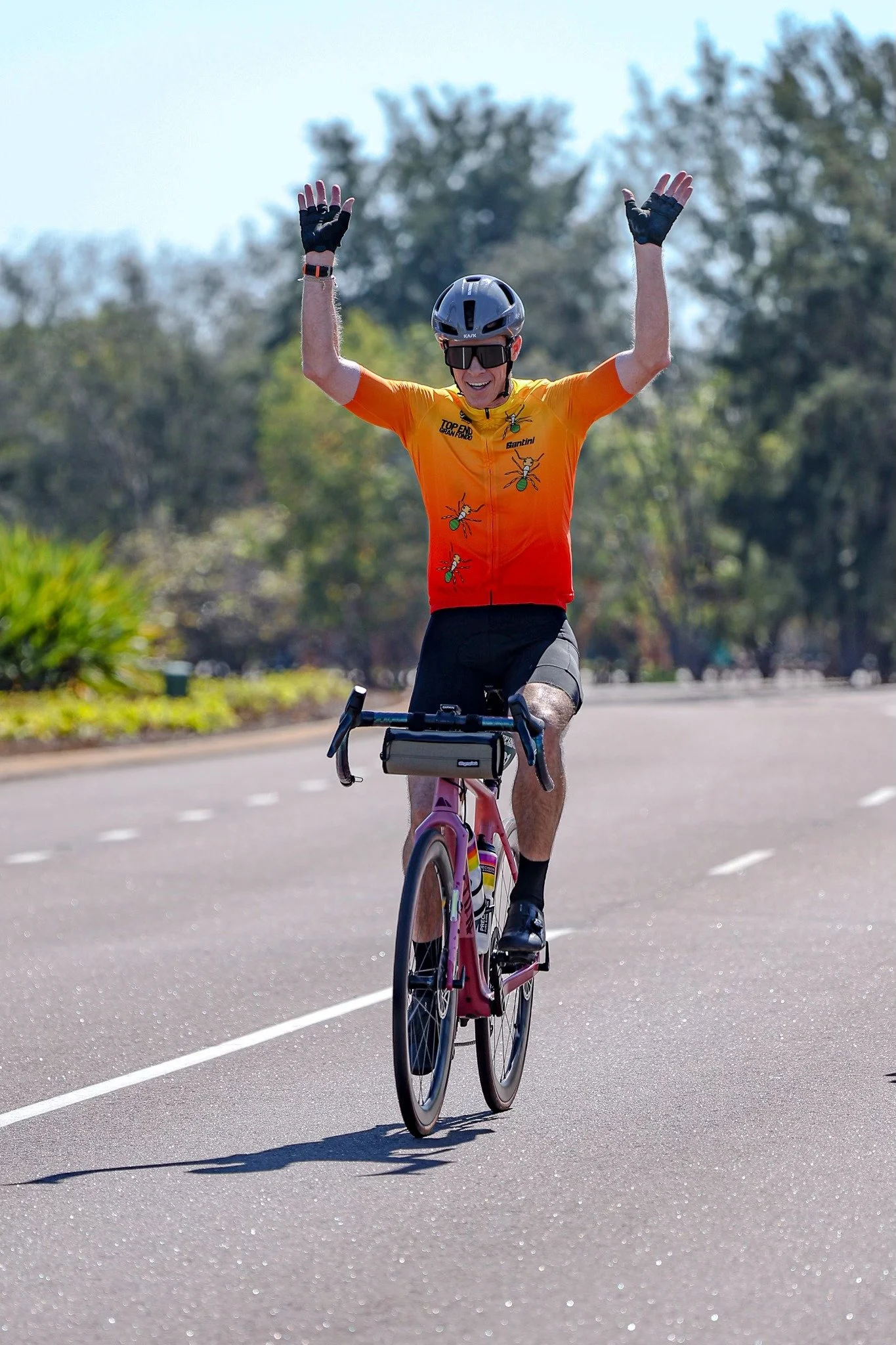 A man riding a pink bicycle on a paved road with greenery in the background, raising both hands in celebration.
