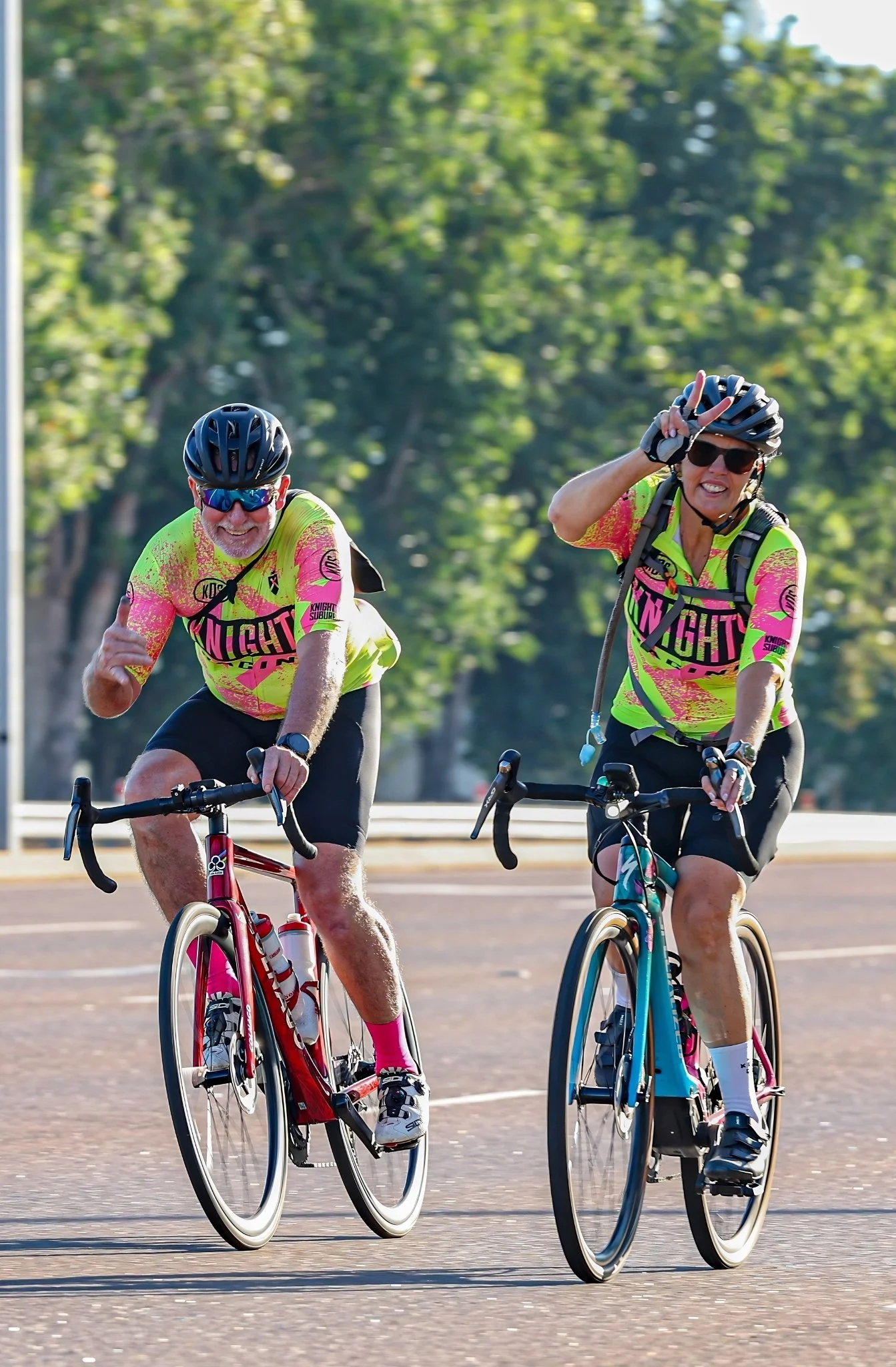 Two cyclists riding on a road, wearing helmets and bright neon shirts, surrounded by trees.