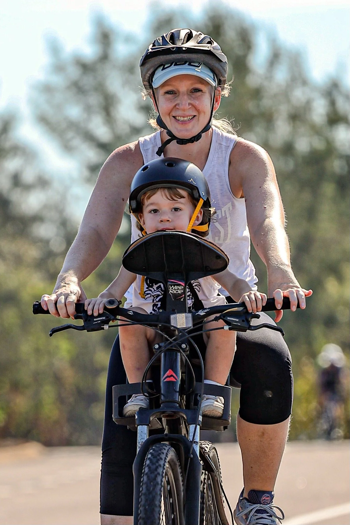 A woman and young boy riding a bicycle outdoors, both wearing helmets and smiling, with a blurred background of trees and sky.