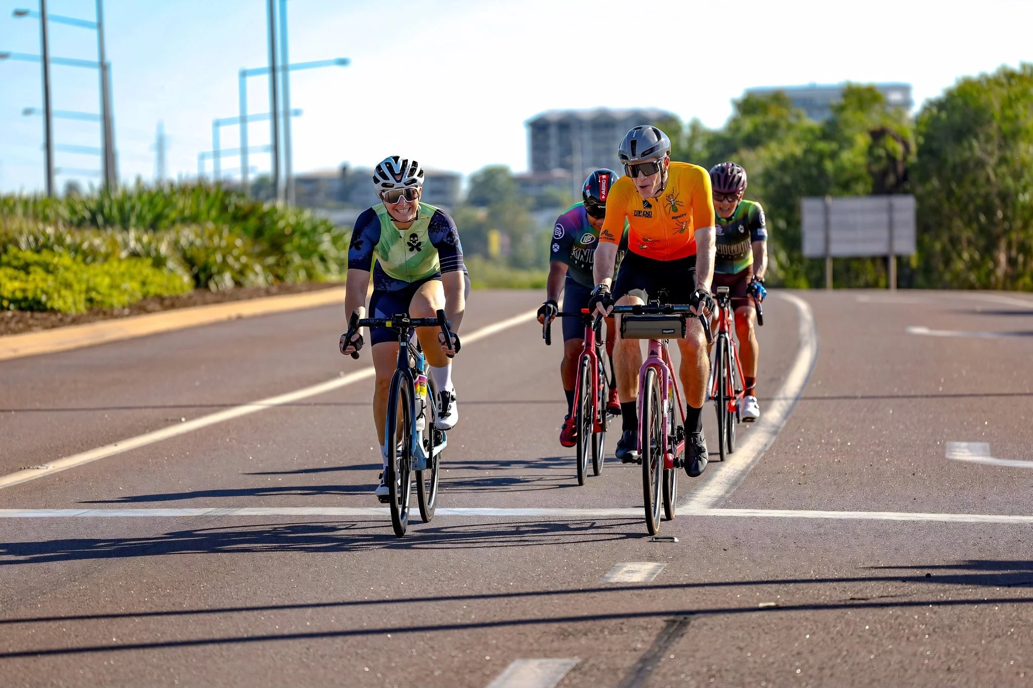 A group of five cyclists riding on a paved road during daytime, wearing colorful cycling gear and helmets, with greenery and buildings in the background.