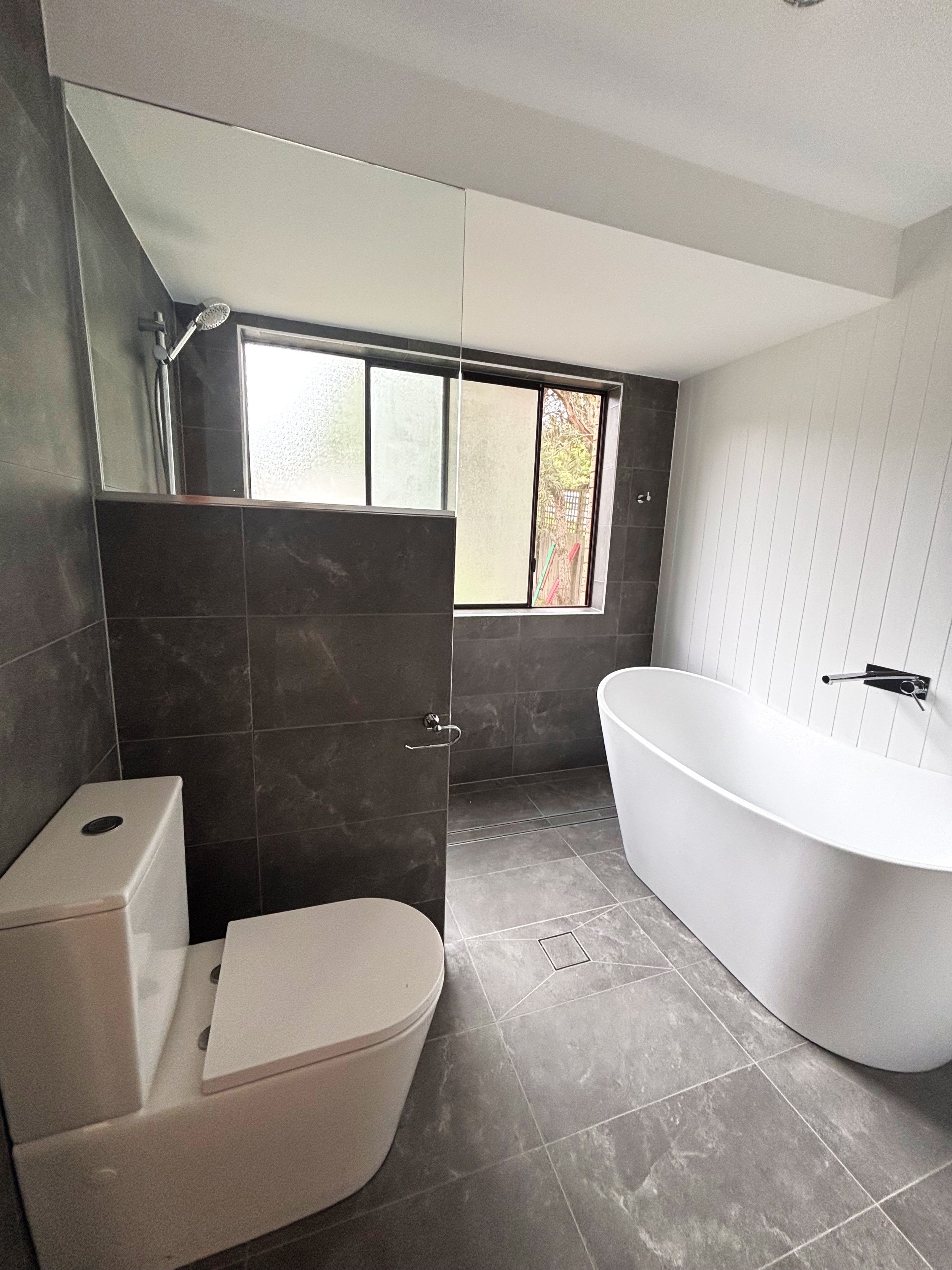 Modern bathroom with dark tiled walls and floor, a white toilet, a freestanding white bathtub, and a frosted glass shower area near a window.