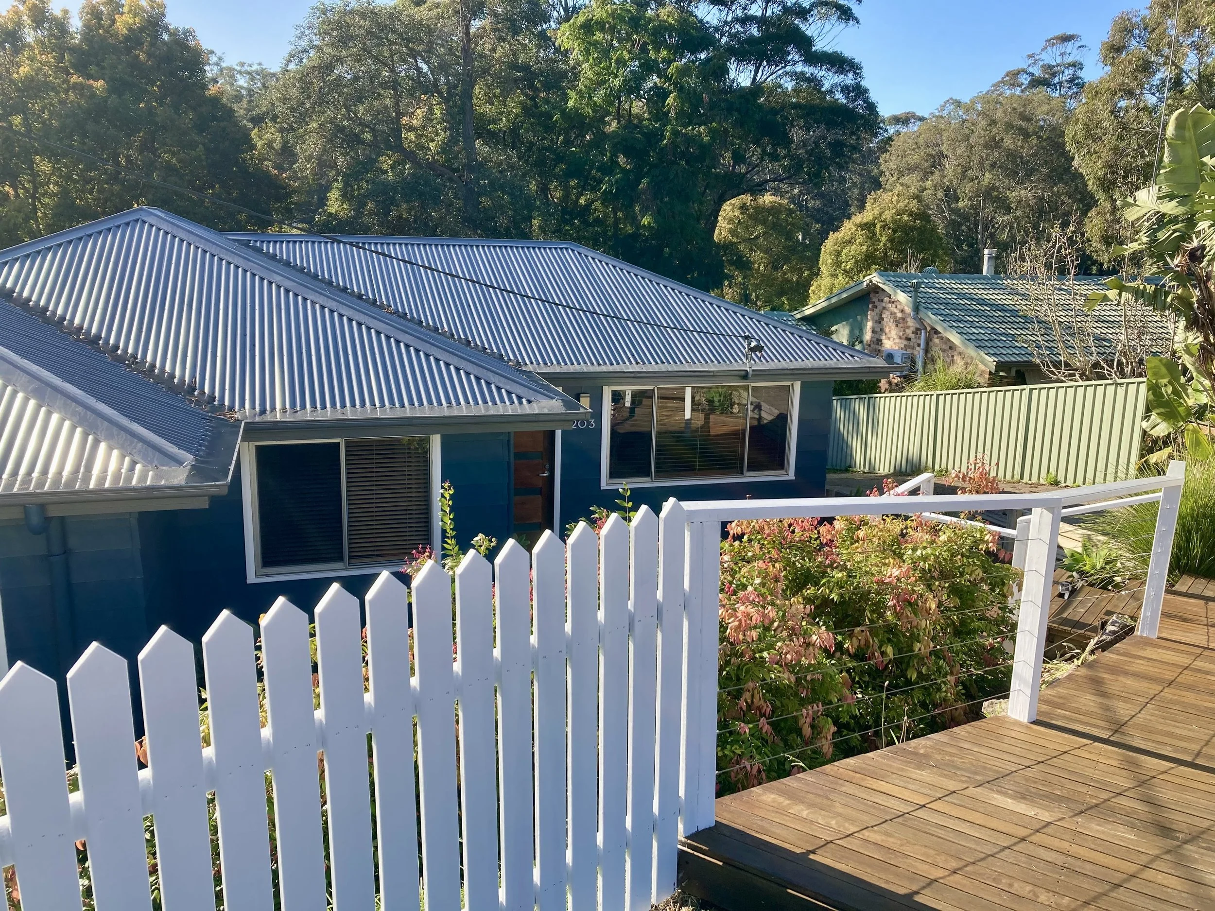 A house with a blue exterior and a metallic roof, surrounded by a white picket fence and a wooden deck, with trees and greenery in the background.