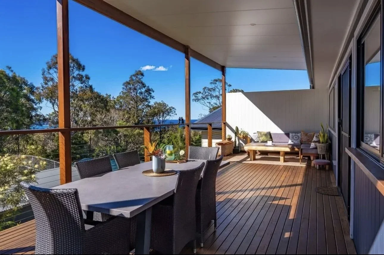 Outdoor balcony with wooden floor, dining area with six black chairs and a gray table, and a seating area with a couch and potted plants under a blue sky with trees in the background.