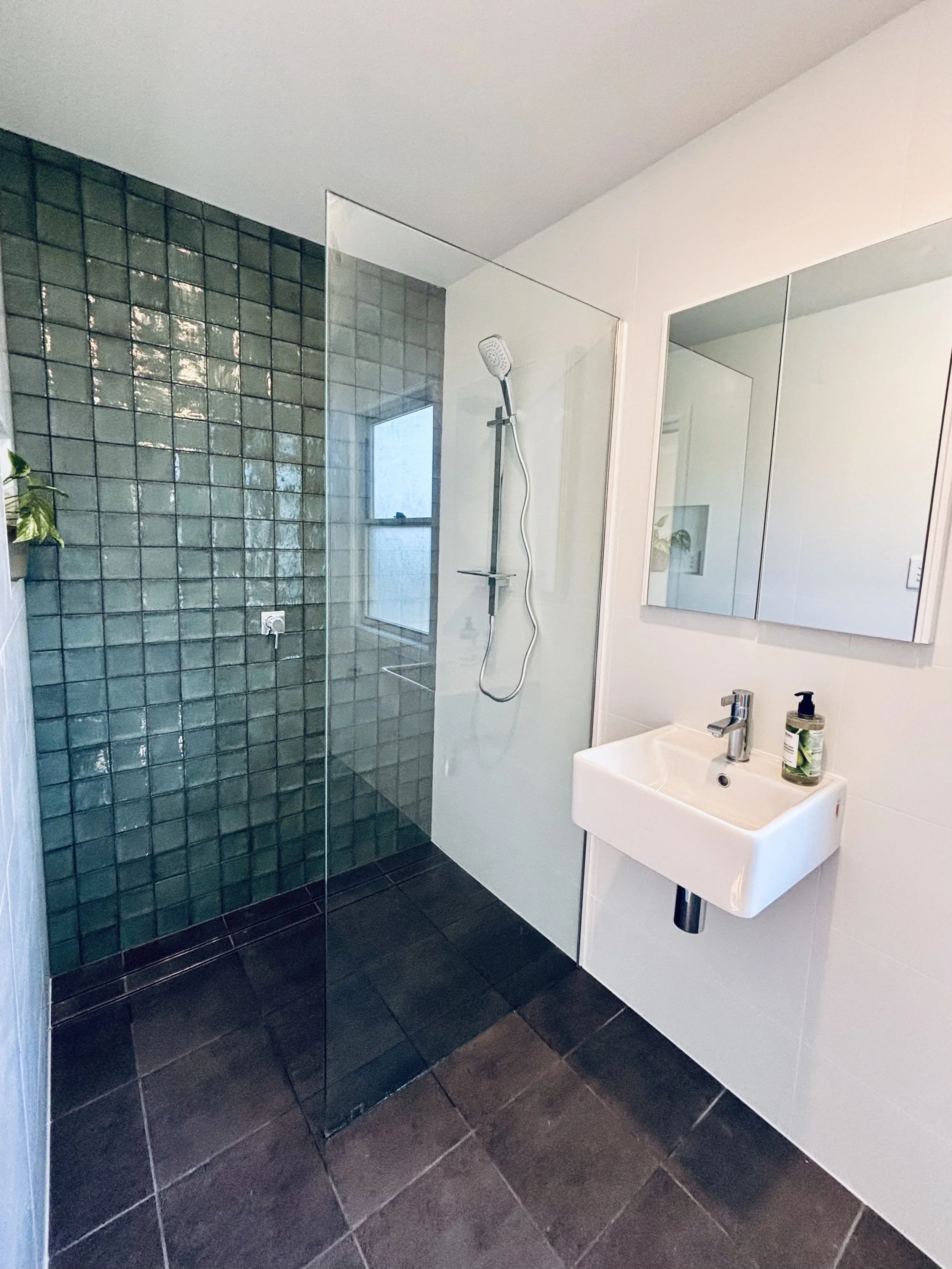 Modern bathroom with a walk-in shower with dark green tiles, a white sink with a mirror cabinet, and a bottle of soap on the sink.