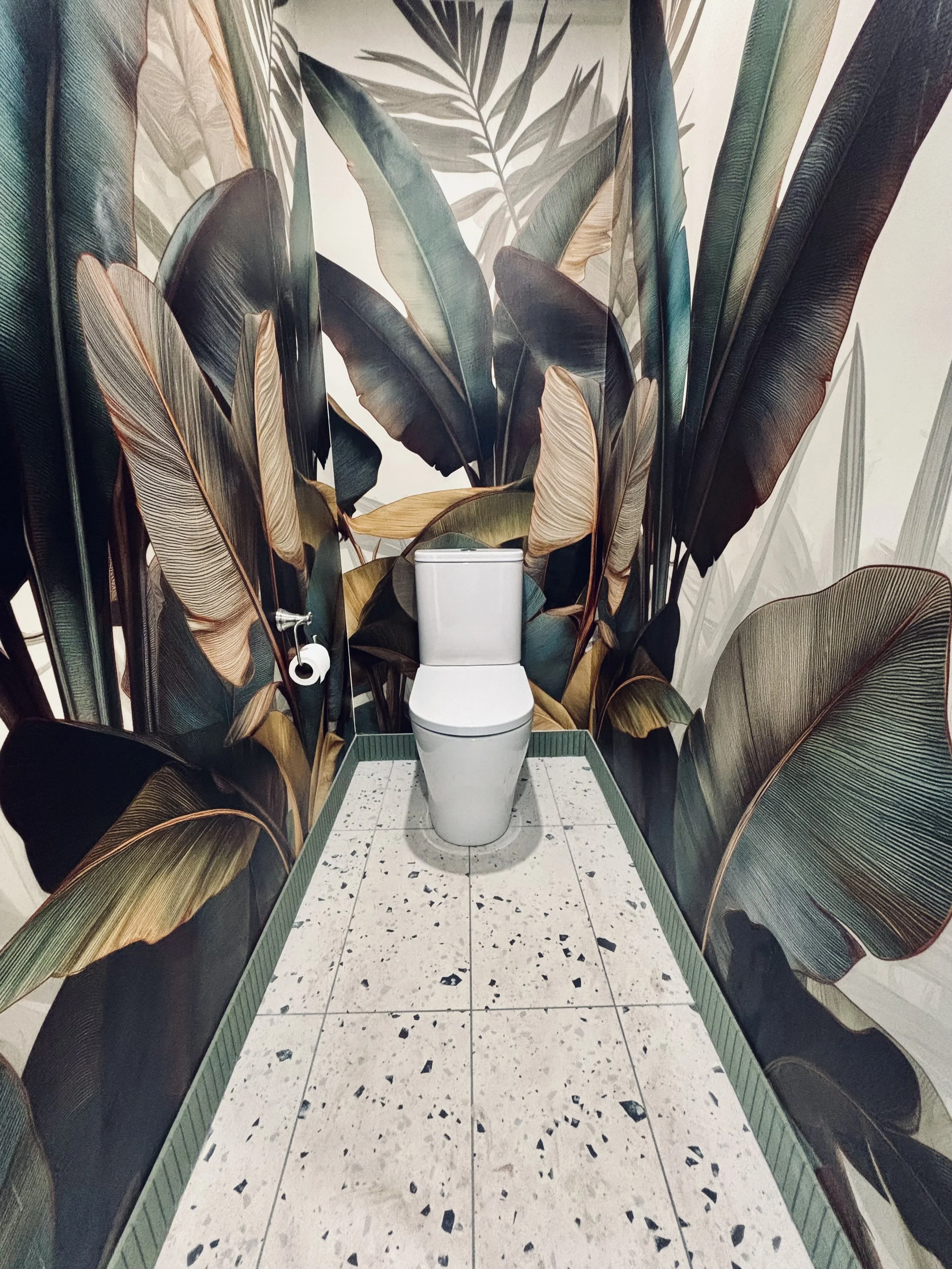 Toilet in a bathroom with tropical leaf-themed wallpaper and terrazzo-style floor tiles.