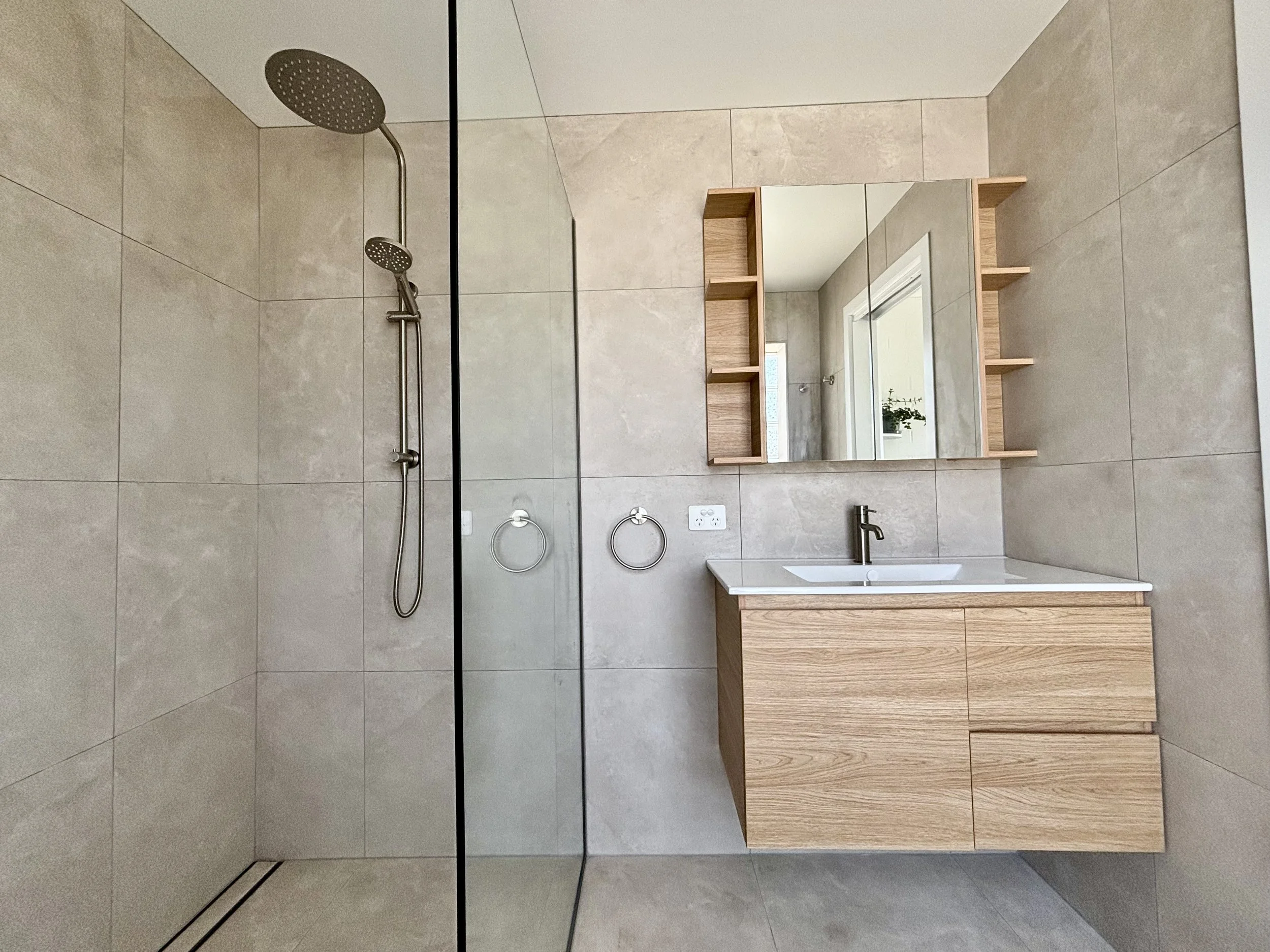 Modern bathroom with a walk-in shower on the left, featuring a rainfall showerhead and handheld shower. The right side has a wooden vanity with a white countertop and an integrated sink, above which is a mirror with built-in shelves. The bathroom wal