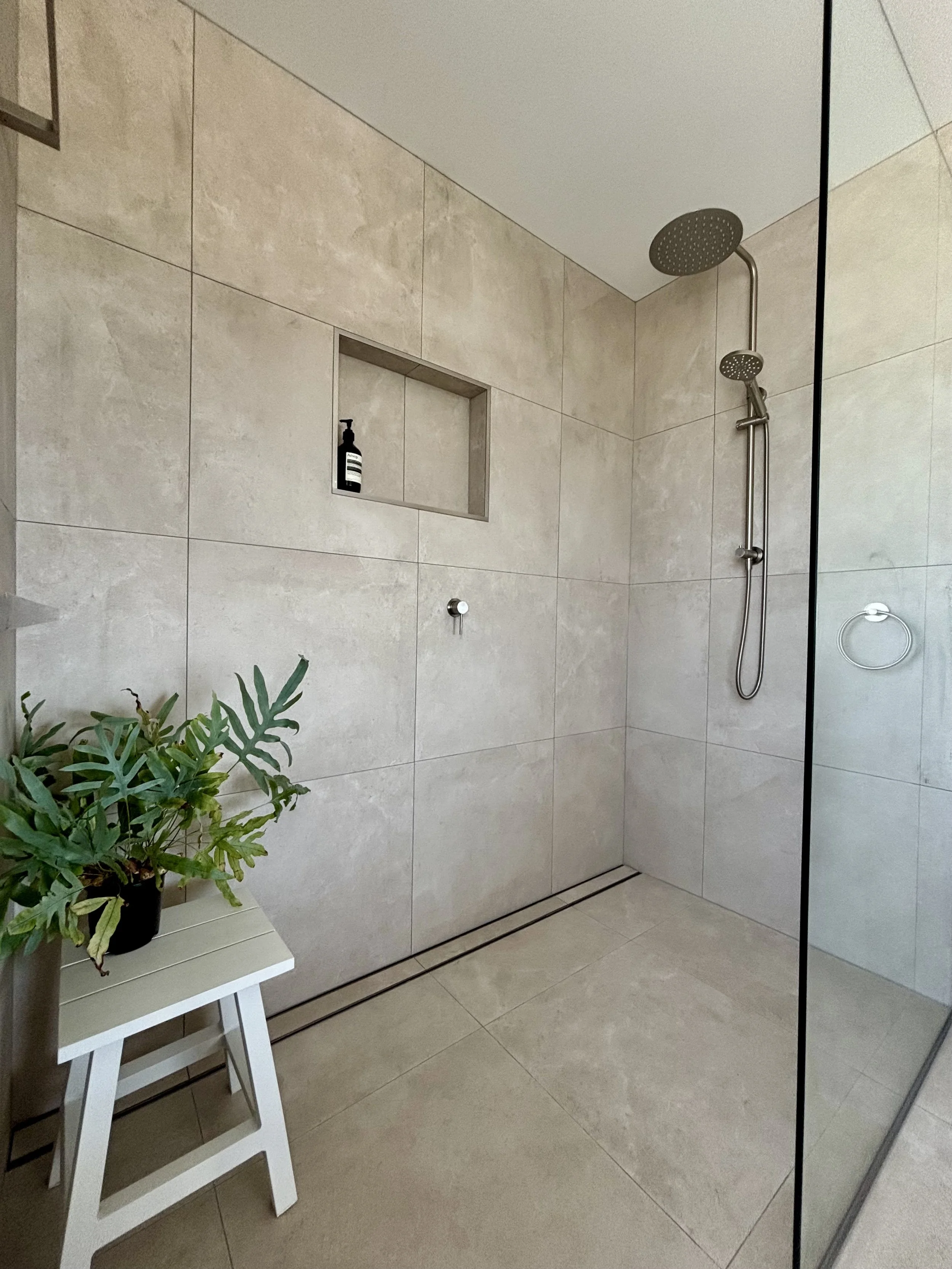 Modern walk-in shower with beige tiles, a built-in niche with bath products, a rain shower head, and a white stool with a green plant placed on it.