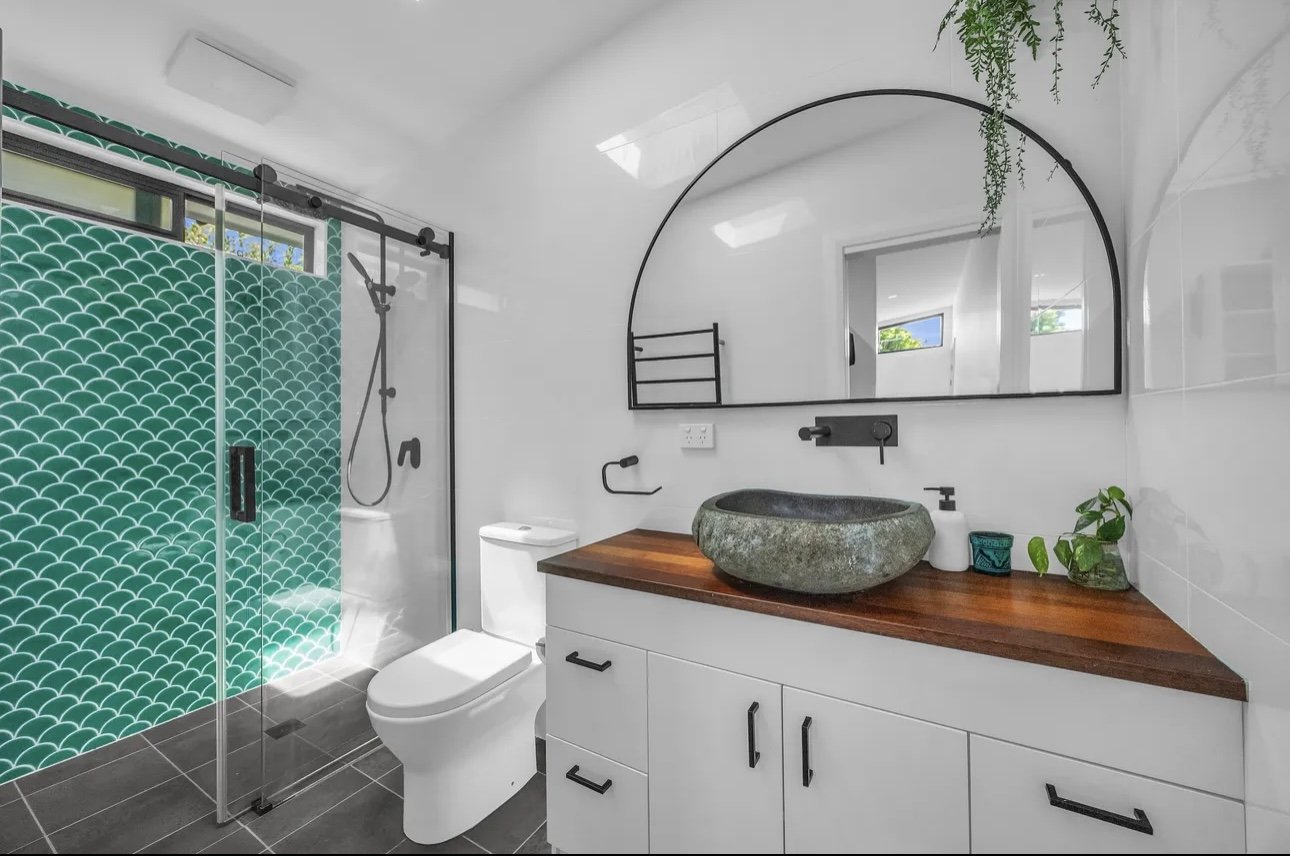 Modern bathroom with a green tiled walk-in shower, white toilet, white vanity with a wooden countertop, round stone sink, black fixtures, large arched mirror, and small plants.