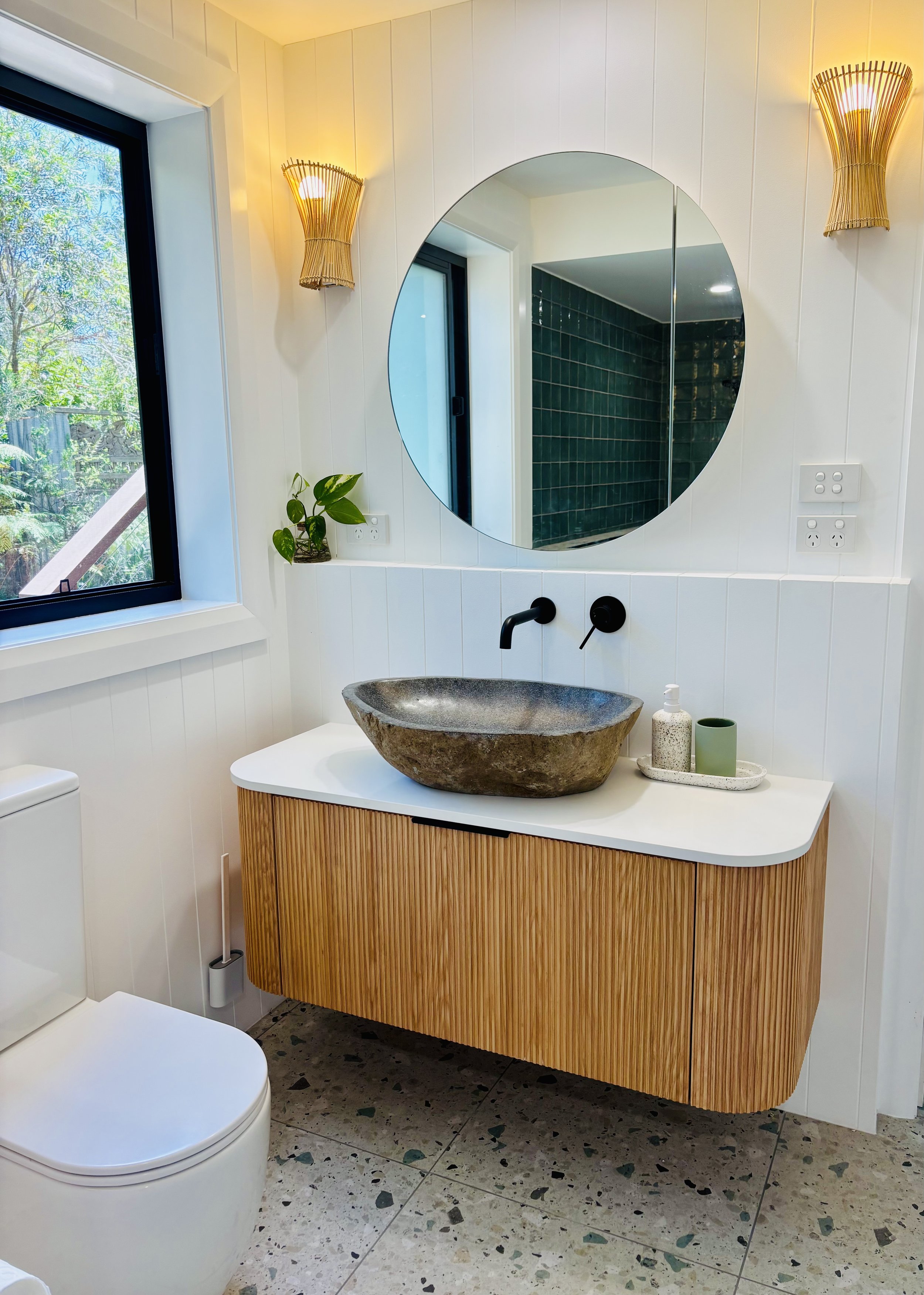 Modern bathroom with a wall-mounted wooden vanity, a stone vessel sink, black wall-mounted faucet, a round mirror, a window with greenery outside, and two wall sconces with woven shades.