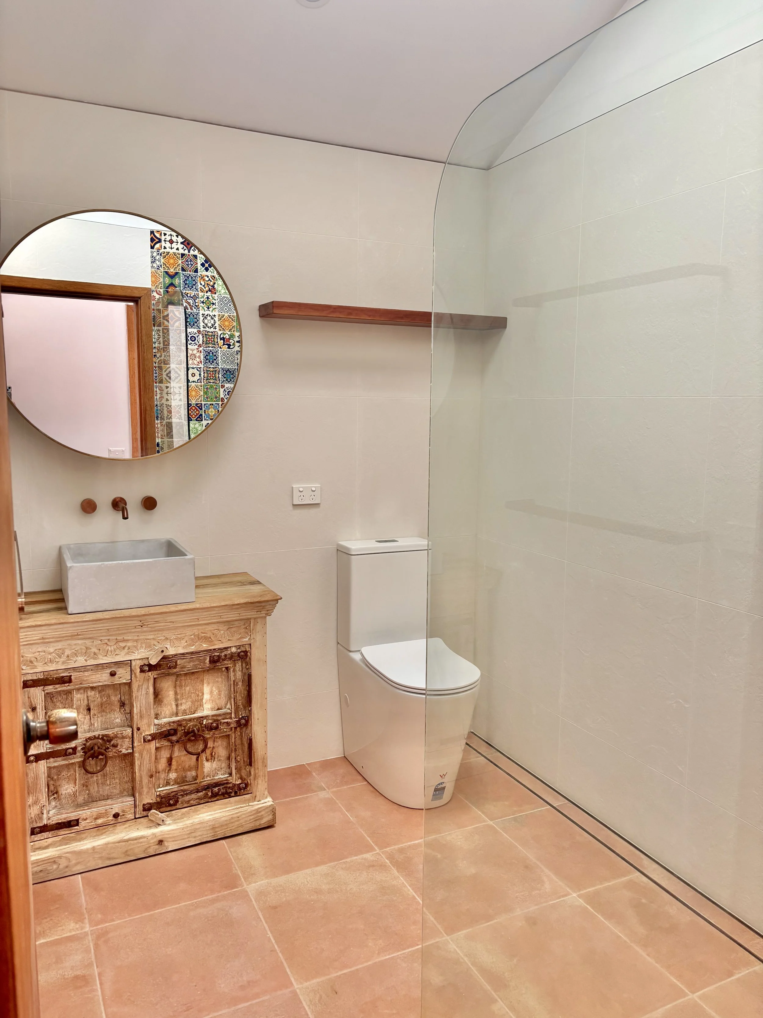 A bathroom with a wooden vanity, a concrete vessel sink, wall-mounted faucet, round mirror, a toilet, two wooden shelves, and a curved glass shower screen, with beige walls and terracotta floor tiles.