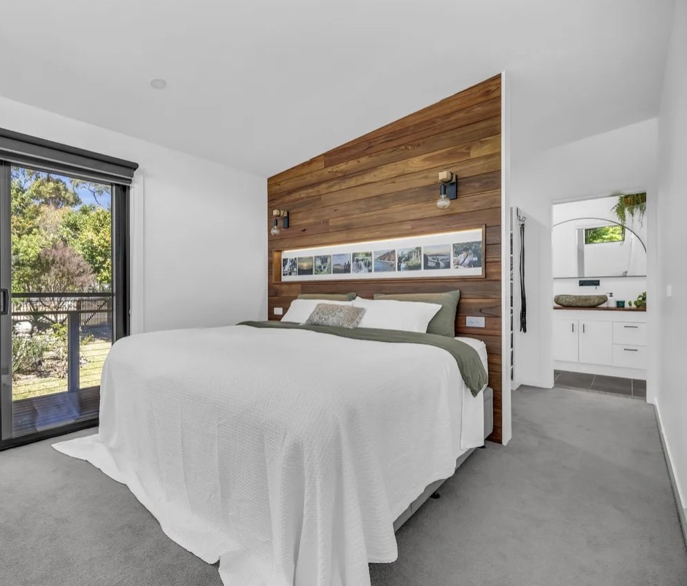 A modern bedroom with a white bed, green bedding, and a wooden accent wall above the headboard. There is a sliding glass door leading outside and a small area with a sink and mirror visible through an arched doorway.