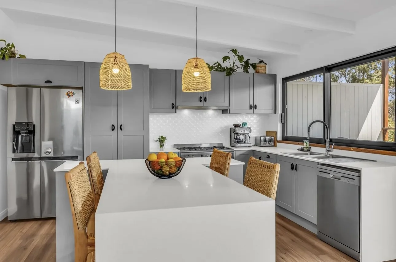 Modern kitchen with gray cabinets, white countertops, stainless steel appliances, a white island with a fruit bowl, bamboo pendant lights, and large windows with a view of trees outside.