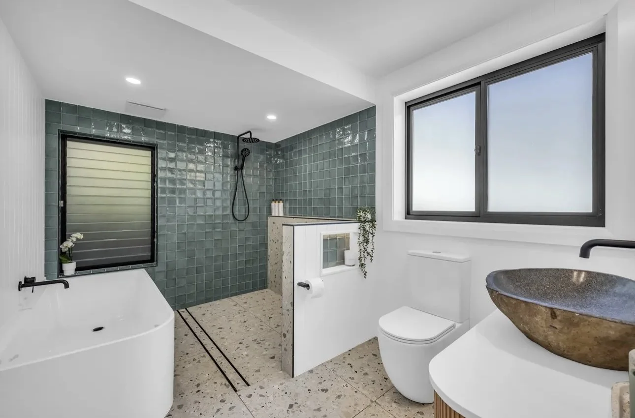 Modern bathroom with a walk-in shower with green tiles, a large window, a white toilet, a black bowl sink, a bathtub, and minimal decor.