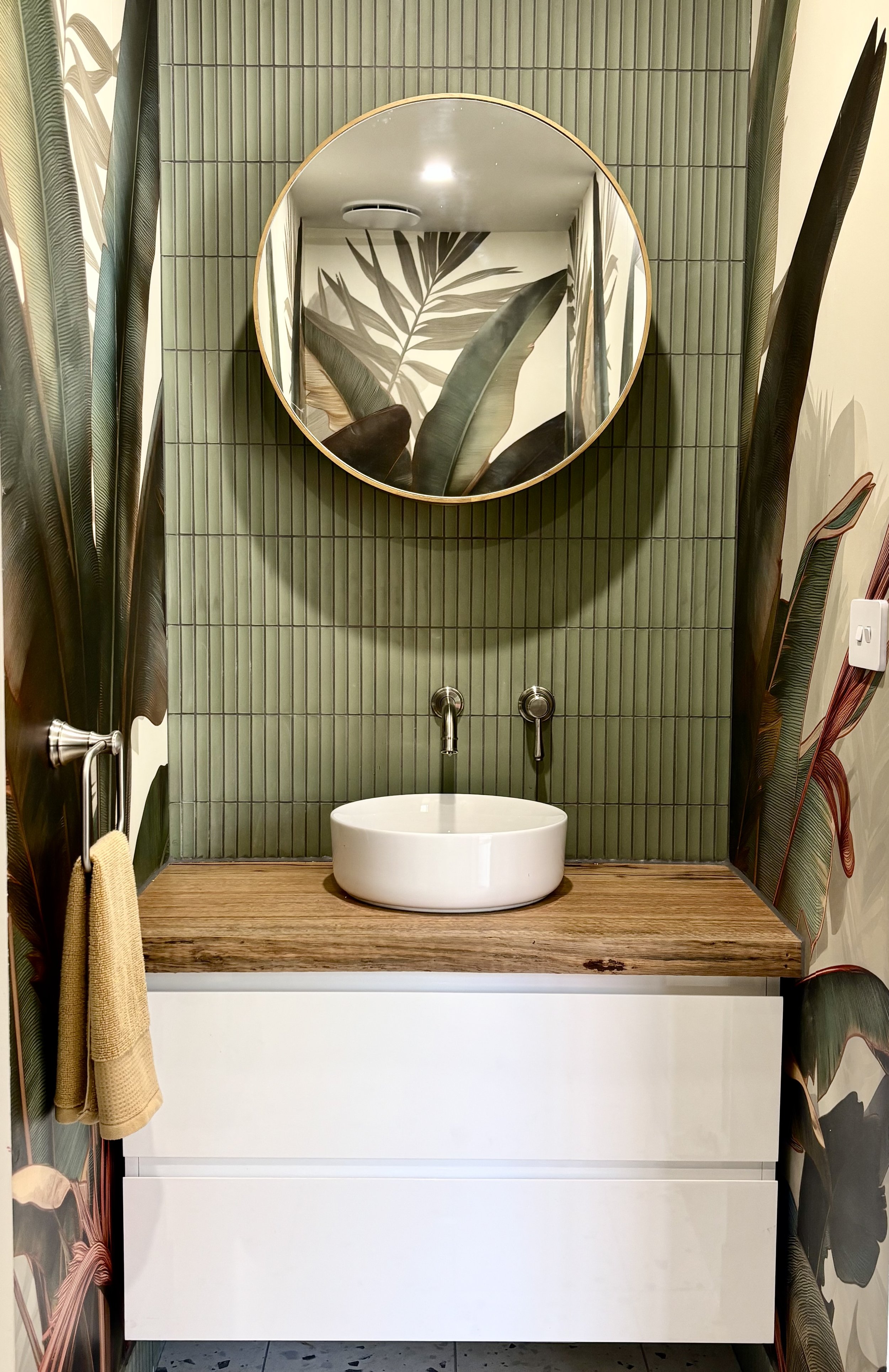 A bathroom with green tile wall, white cabinet, and wooden countertop holding a round white vessel sink. A round mirror with a wooden frame reflects tropical leaf wallpaper, and large tropical leaves are on the walls.