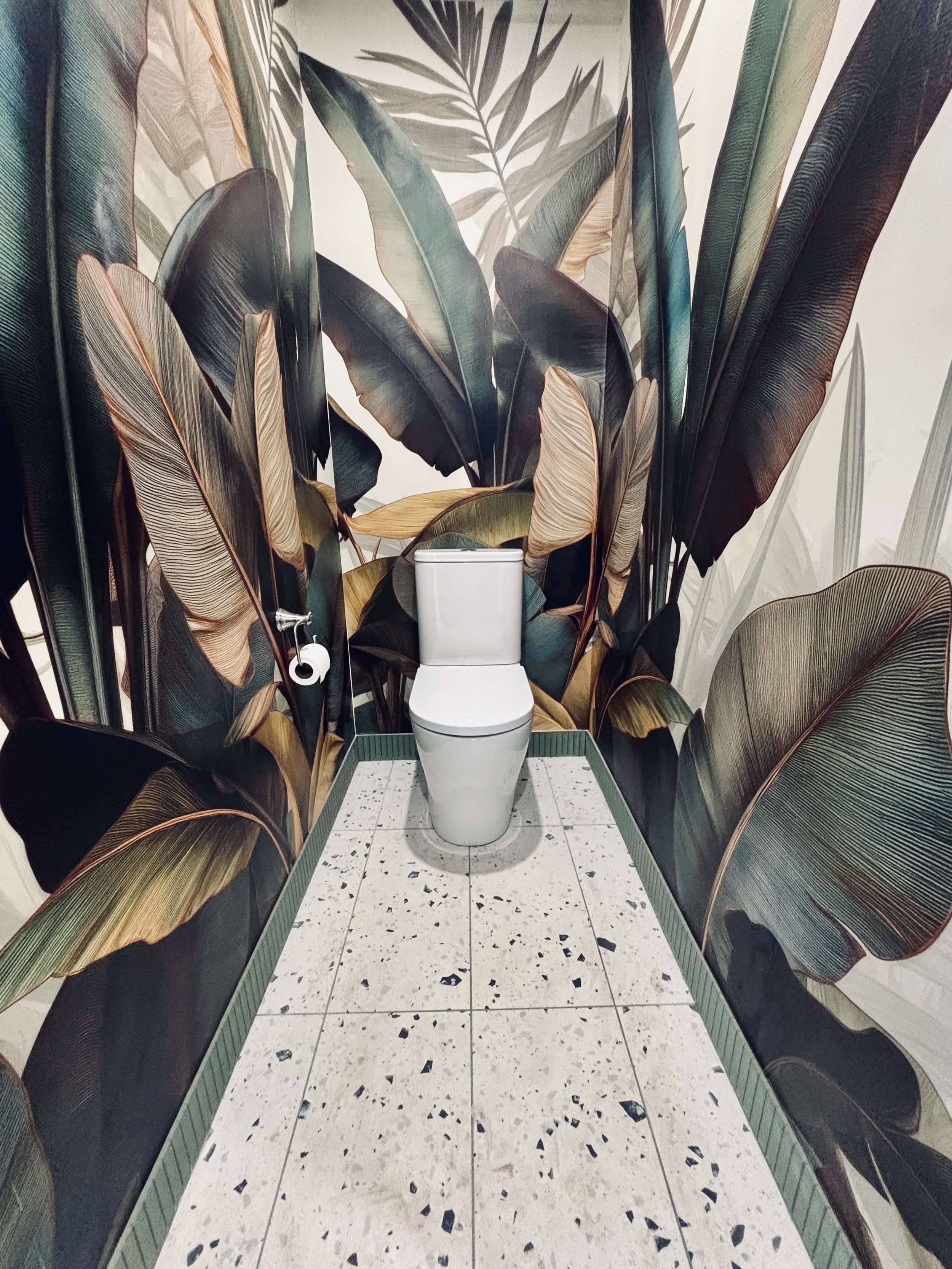 A modern toilet in a small room with tropical leaf-patterned wallpaper and terrazzo tile flooring.
