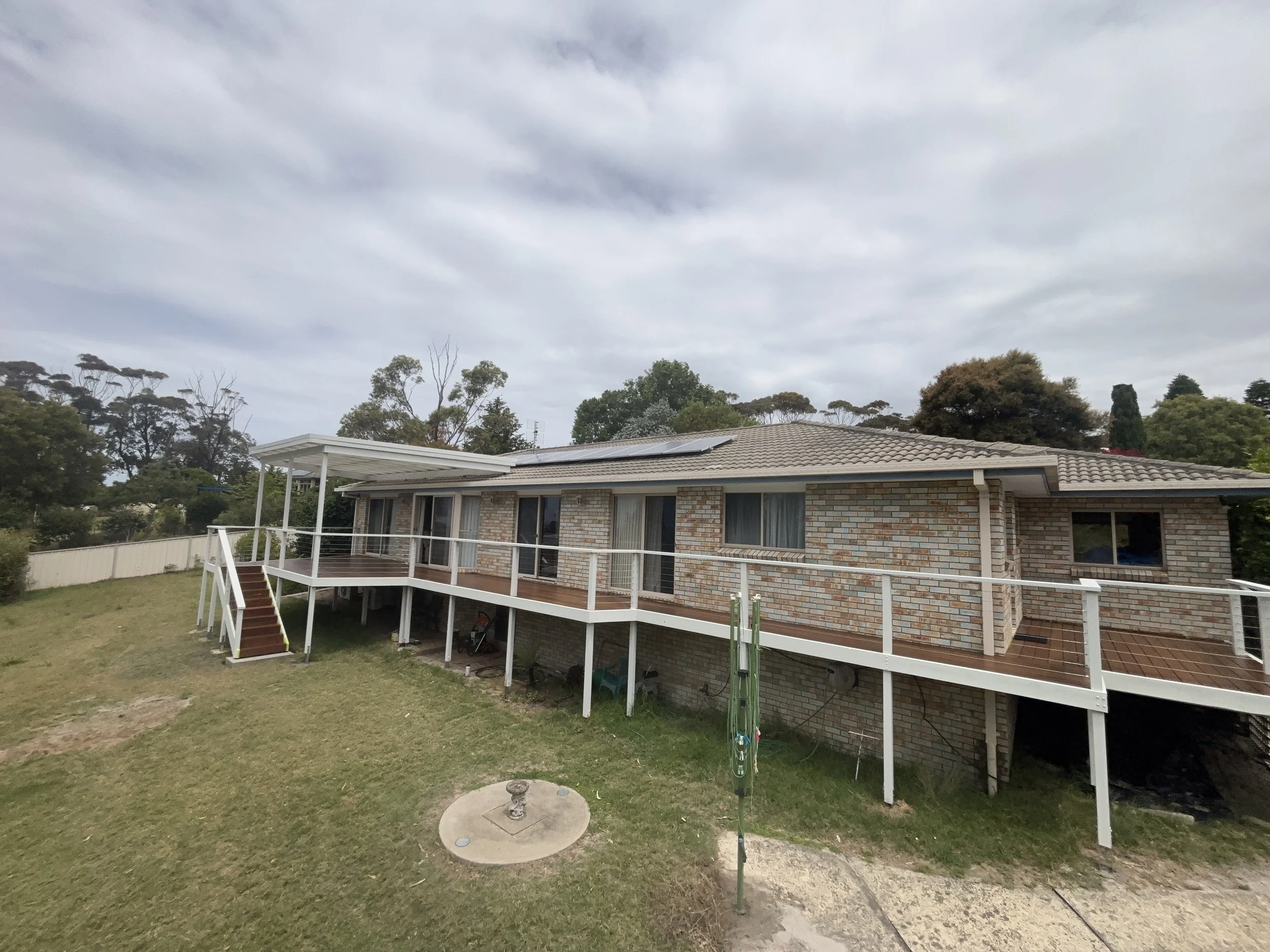 A brick house on a raised foundation with a large wooden deck and white railing, surrounded by a grassy yard and trees in the background under a cloudy sky.
