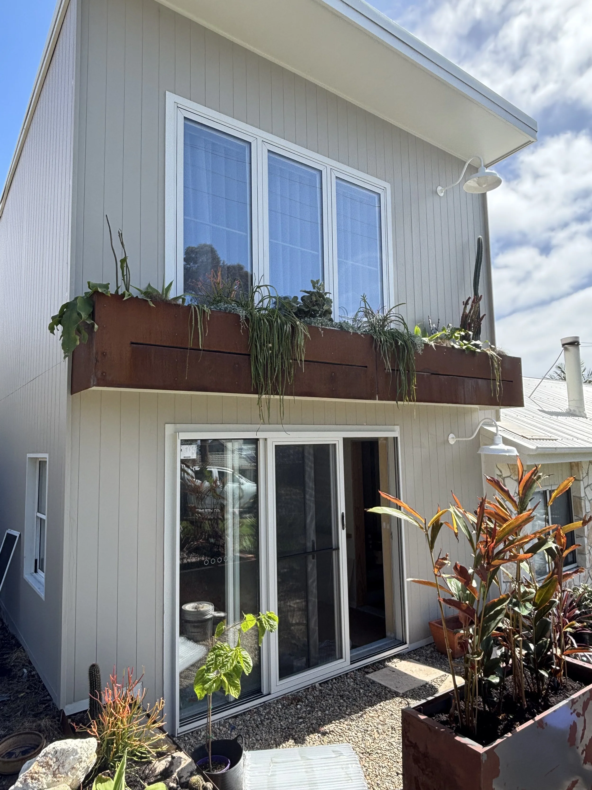 The image shows the rear view of a two-story house with white vertical siding, a large window on the upper floor with curtains, and a sliding glass door on the ground floor. There's a balcony with a rusty metal railing and plants, including cacti and