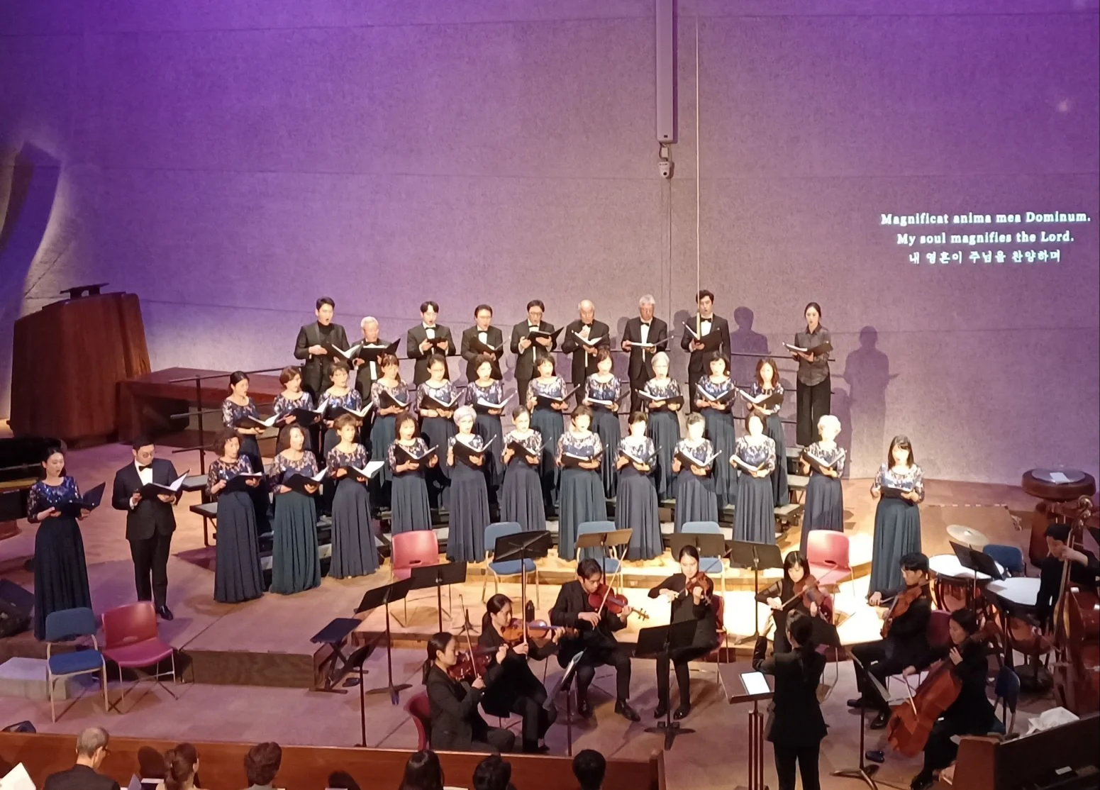 A choir of adult men and women dressed in formal black and blue gowns singing on stage, accompanied by a string orchestra with musicians playing violins, viola, and cello in front of a purple backdrop, with song lyrics projected on the wall reading '