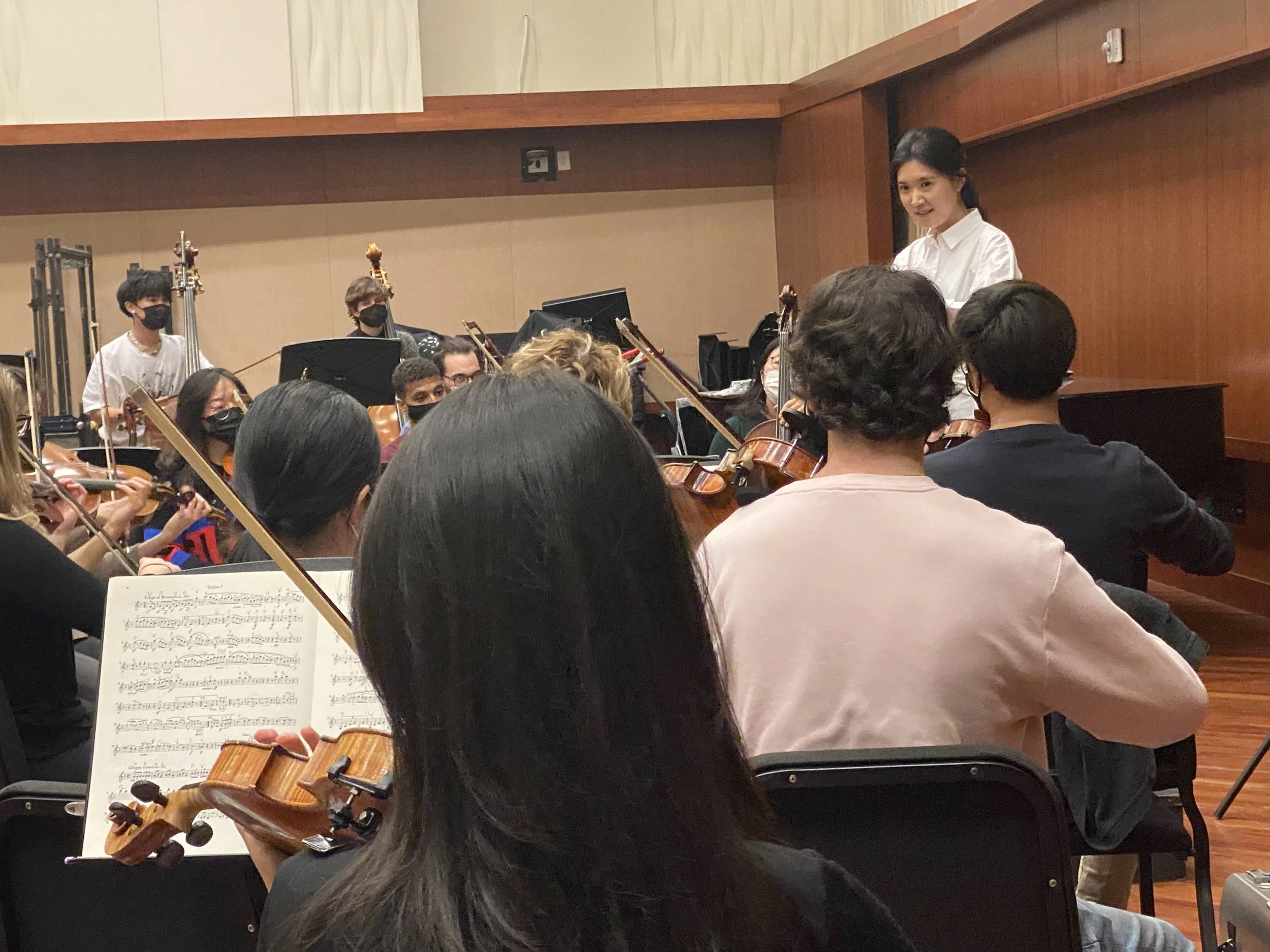 Orchestra rehearsing in a concert hall, with a conductor smiling at the musicians, who are playing string instruments and wearing black masks.