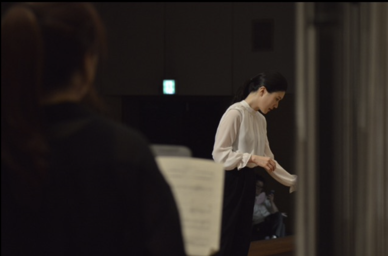 A woman in a white blouse and black skirt preparing for a performance in a dimly lit room, with a person in black in the foreground holding a sheet of music.