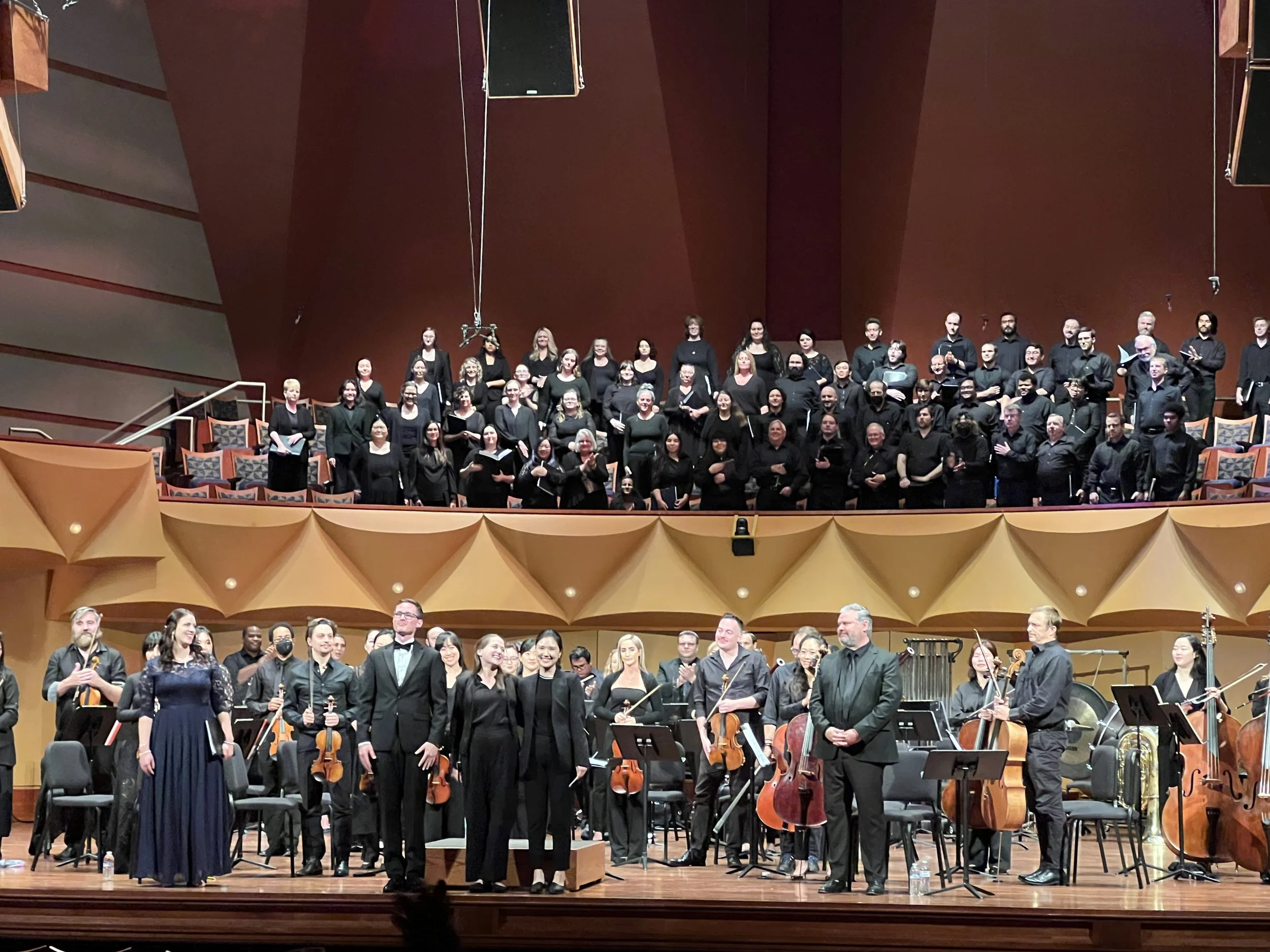 A large choir and an orchestra on stage after a performance in a concert hall. The choir stands on the upper level, dressed in black, and the orchestra members are on the lower level, also in black, holding string instruments.