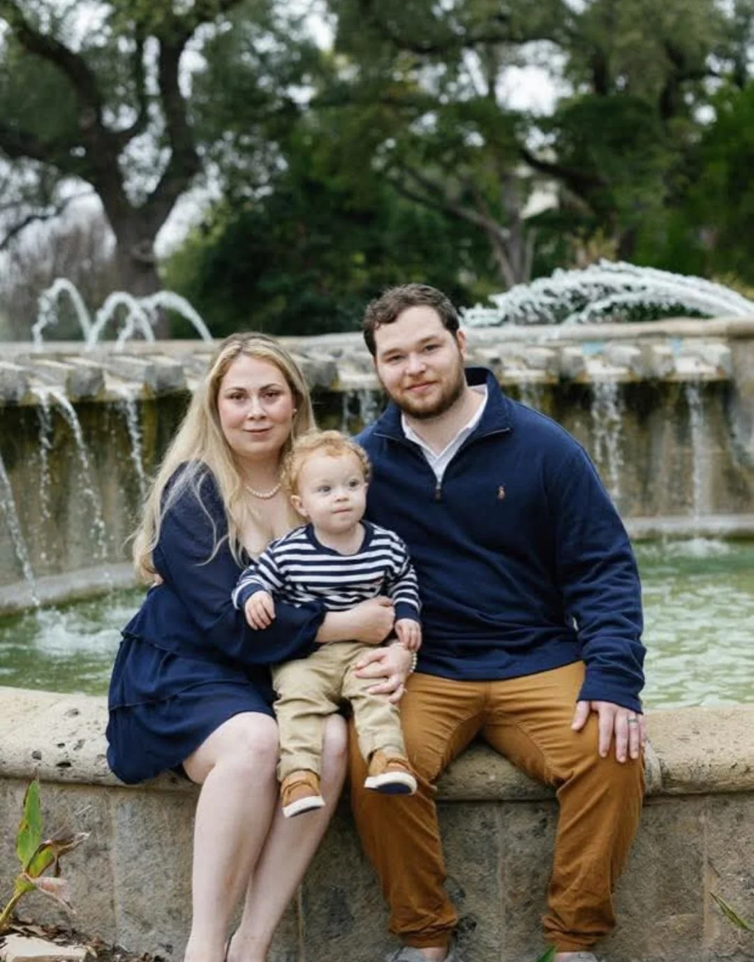 A family of three sitting on a stone ledge in front of a fountain with water spouting, surrounded by trees and greenery.