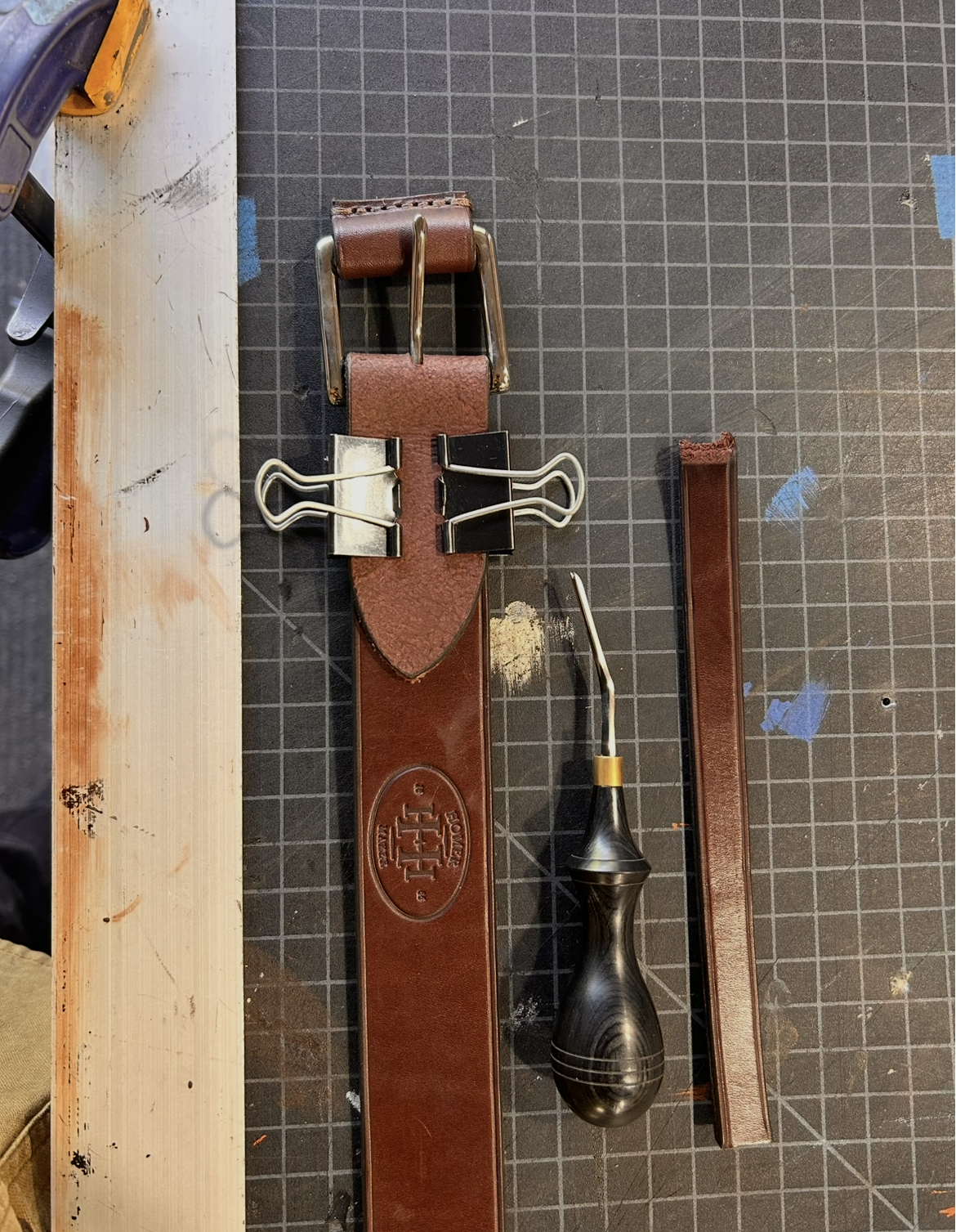 A leather belt with a metal buckle, a saddle hook tool with a black and yellow handle, and a strip of leather on a work table.