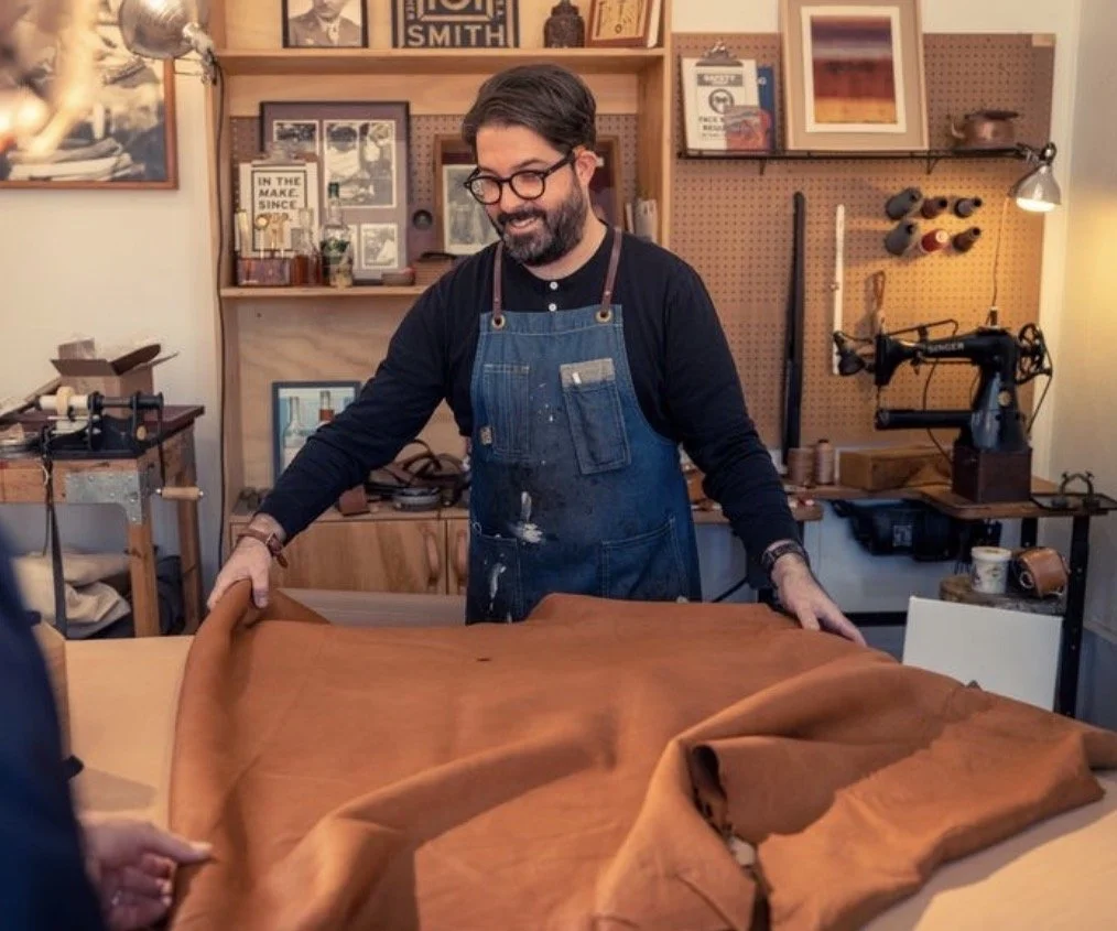 A man with glasses, a beard, and dark hair, wearing a black shirt and blue apron, is smiling while folding a large piece of brown leather in a woodworking or leather workshop.