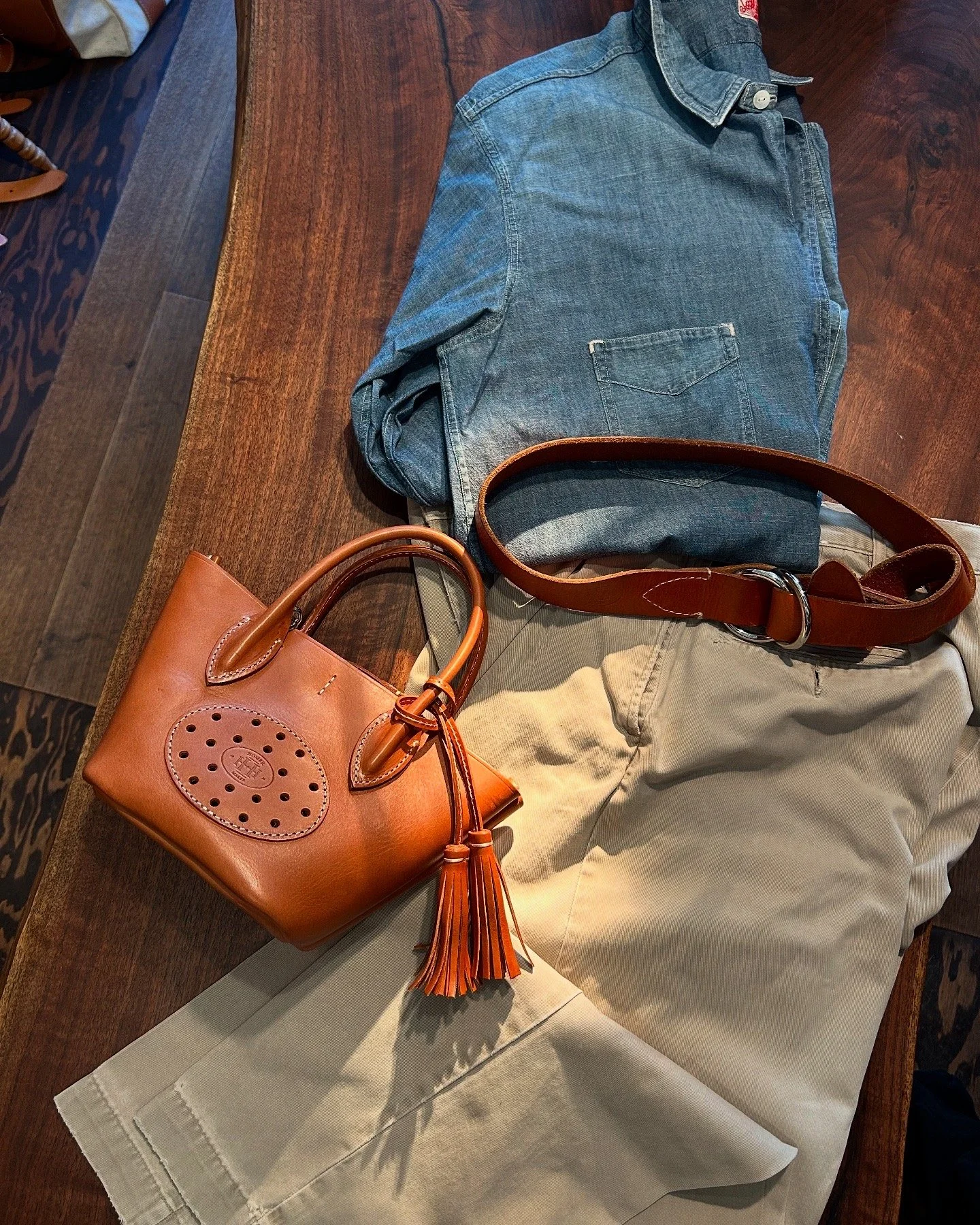 A wooden table with a folded blue denim shirt, a beige pair of pants, a brown leather belt, and a tan leather handbag with a tassel.