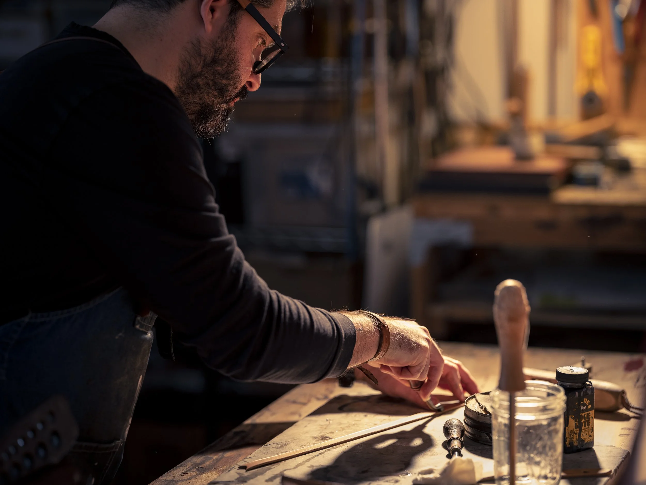 A man wearing glasses and a black shirt working on a craft project at a cluttered workbench, with tools and supplies.