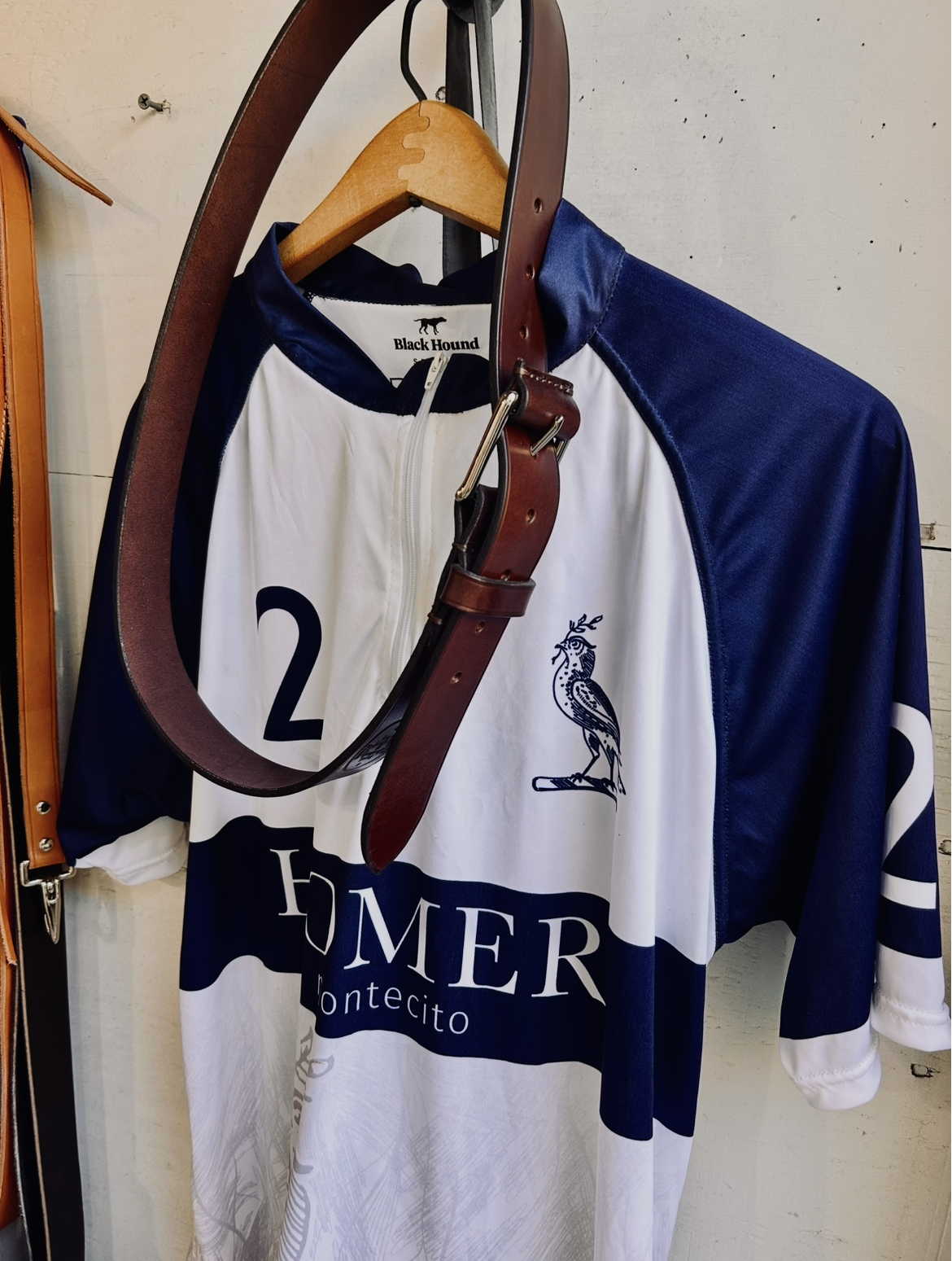 A white and navy blue cycling jersey hanging on a wooden hanger, with a leather belt draped over it. The jersey has a logo of a bird and the words "HOMER Montecito."