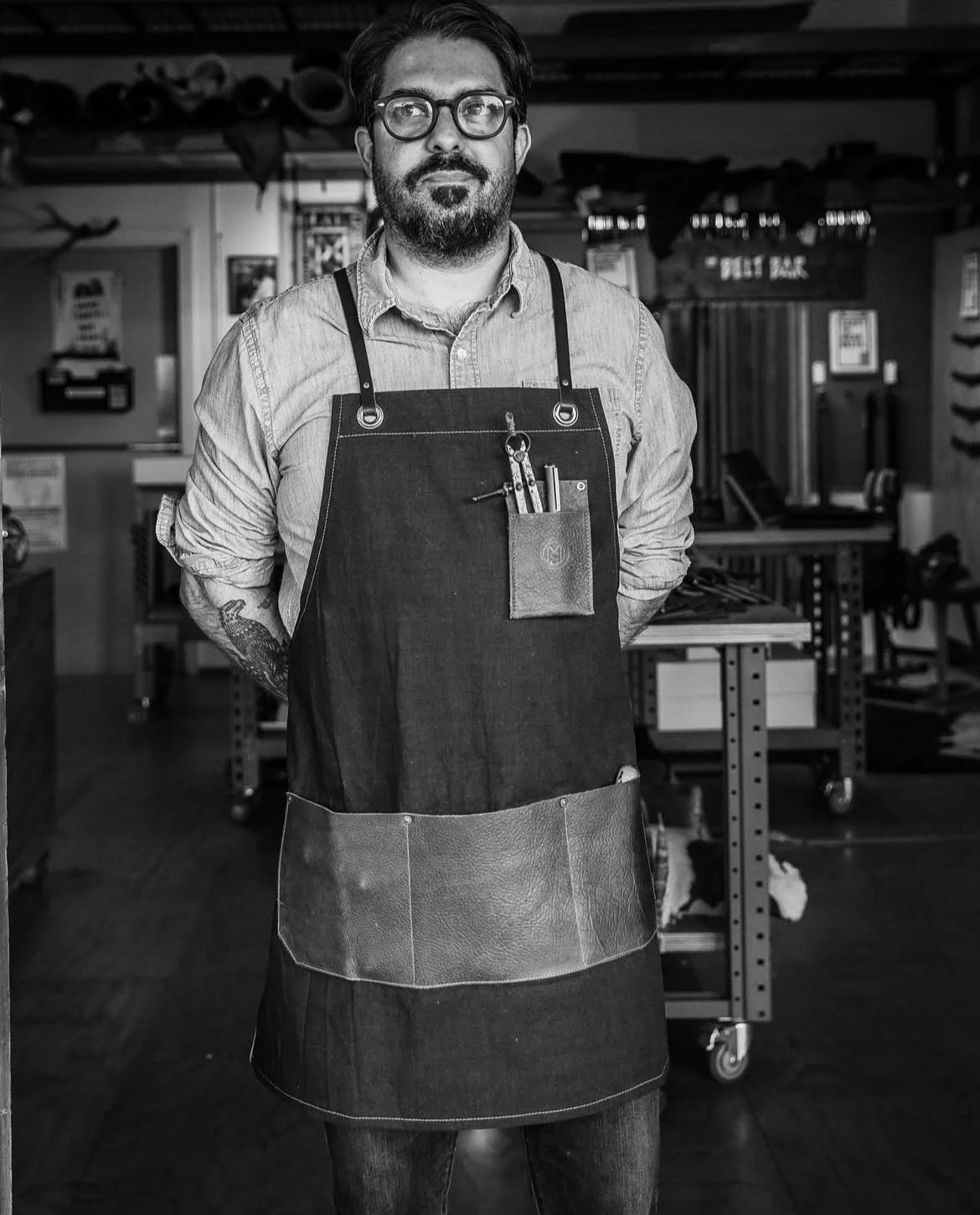 A man with glasses and a beard standing in a workshop, wearing a denim apron with tools in a pocket, in black and white.